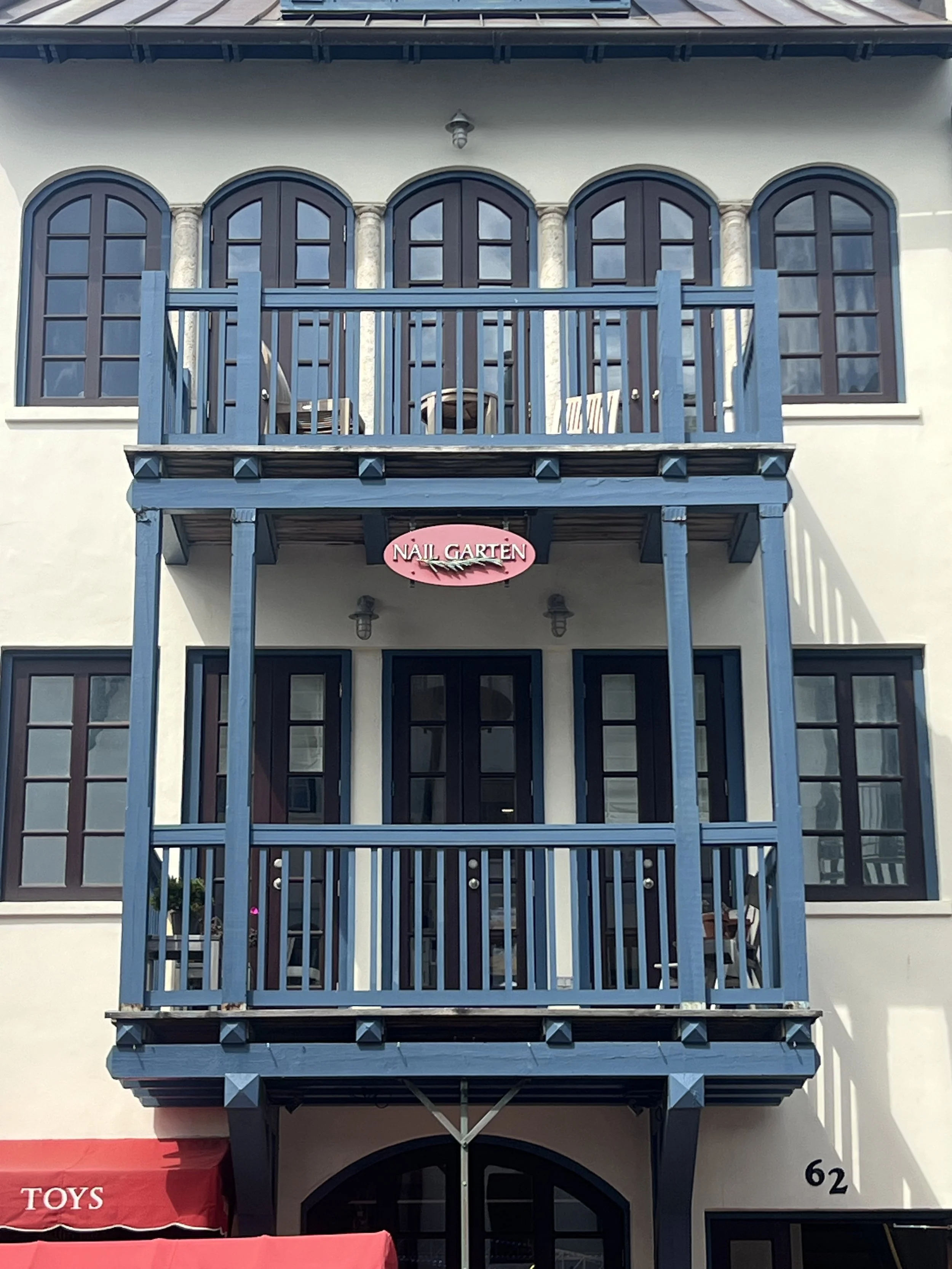 Front of a white building with a blue two-story wooden balcony. The sign on the balcony reads "Nail Garten" in pink. There is a red awning with the word "Toys" at the bottom left.