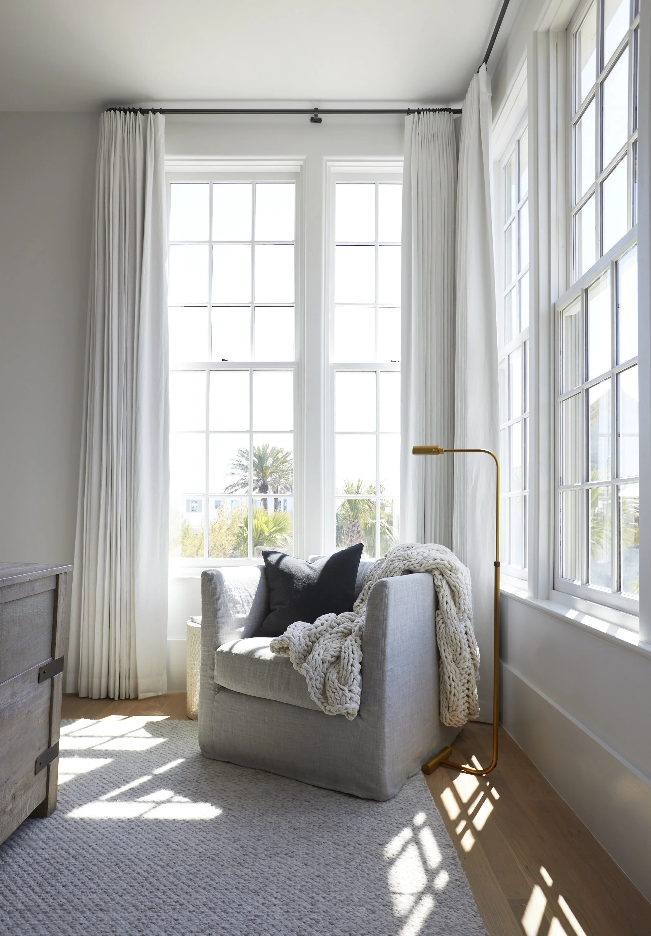 Bright living room corner with large windows, white curtains, a beige armchair with black and beige pillows, a chunky knit throw, a gold floor lamp, and sunlight casting shadows on a neutral area rug.