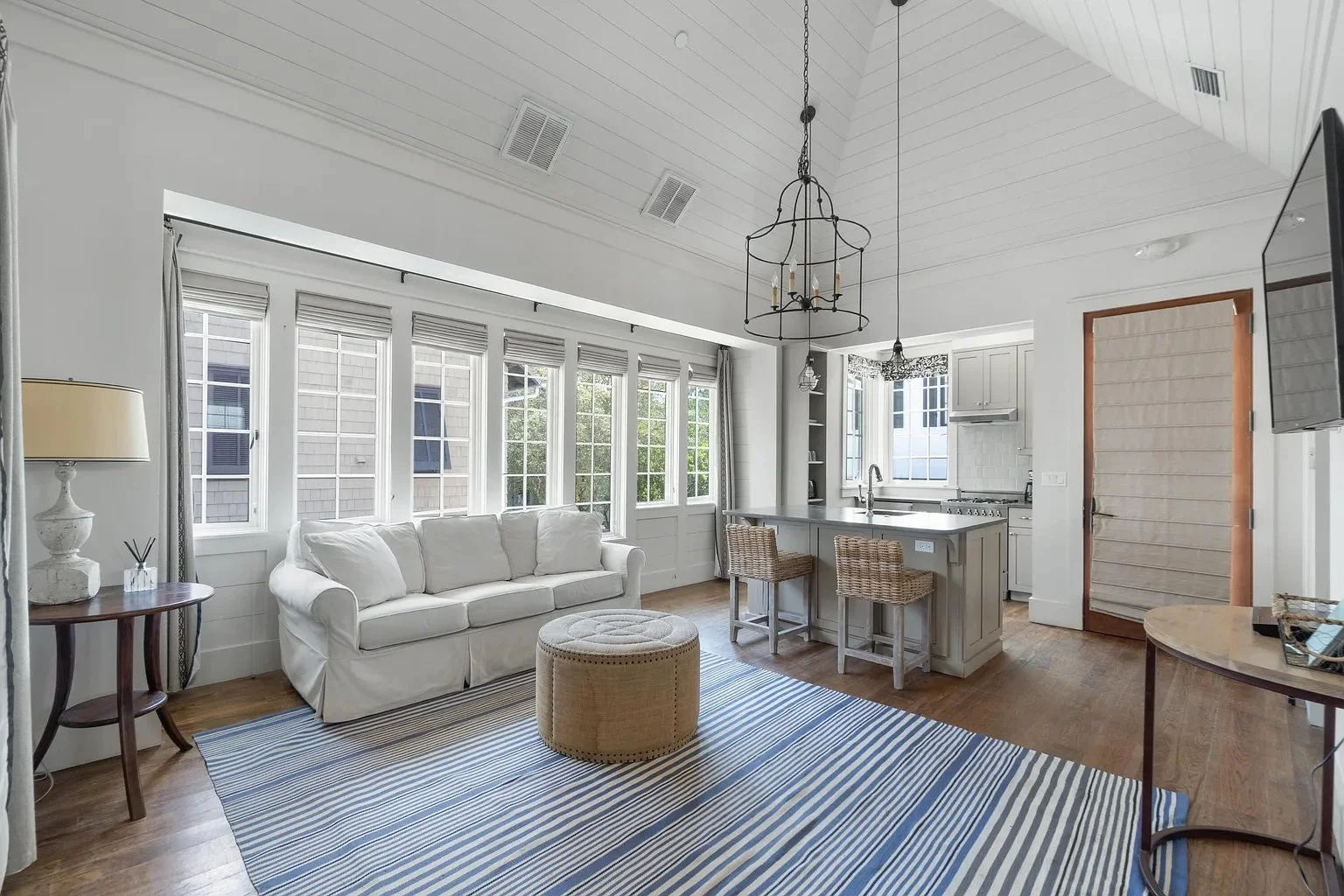 Cozy open-concept living room and kitchen with white walls, large windows, beige sofa, striped blue rug, wooden furniture, and kitchen island with bar stools.