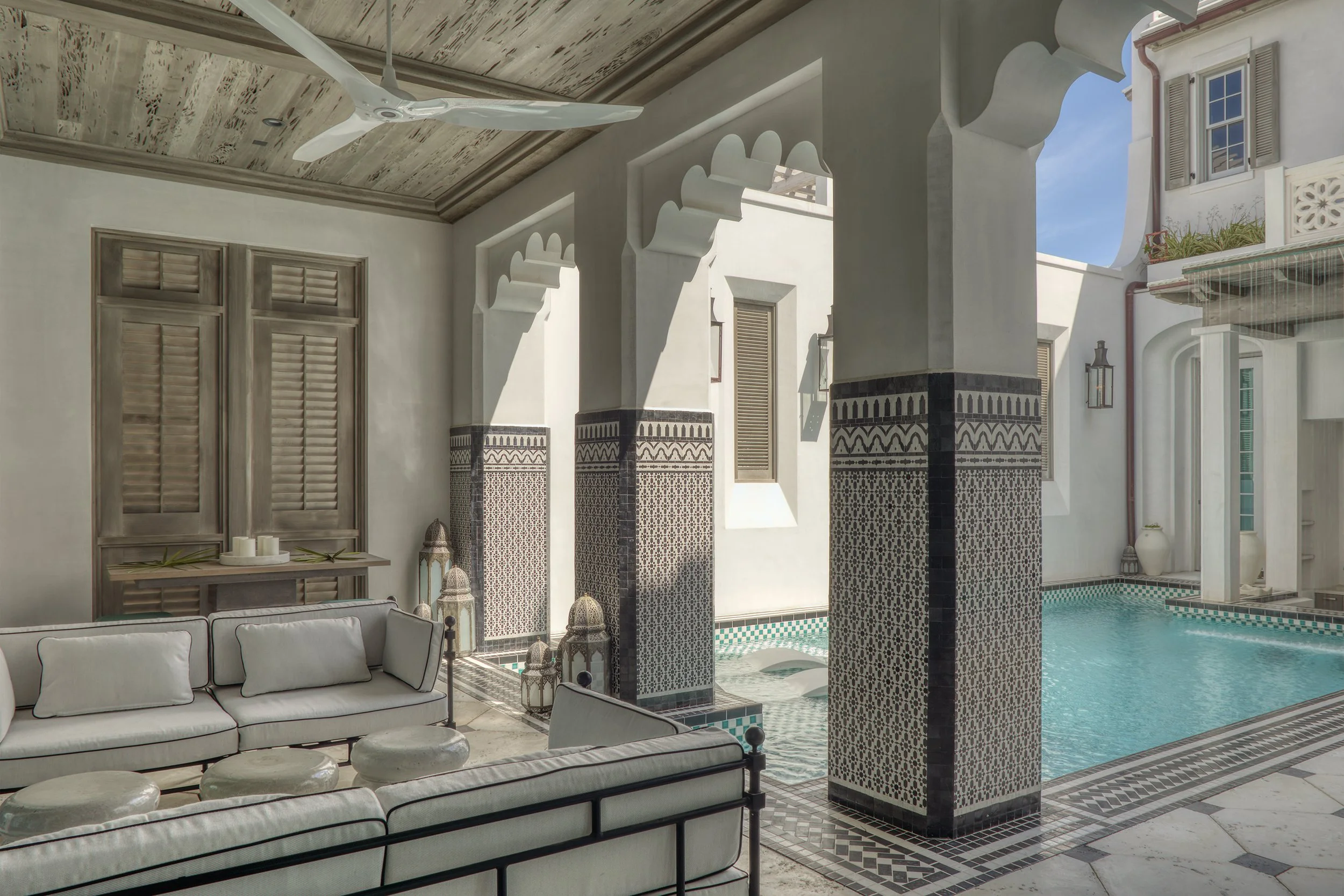 Outdoor pool area with white lounge sofa, decorative lanterns, tiled columns, and a swimming pool under a wooden ceiling fan, with sunlight casting shadows.