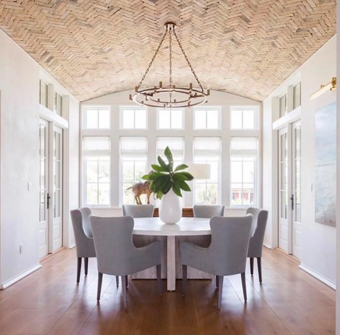 Bright dining room with a round table, six light gray chairs, a white vase with green leaves, large windows, a chandelier, and a brick vaulted ceiling.