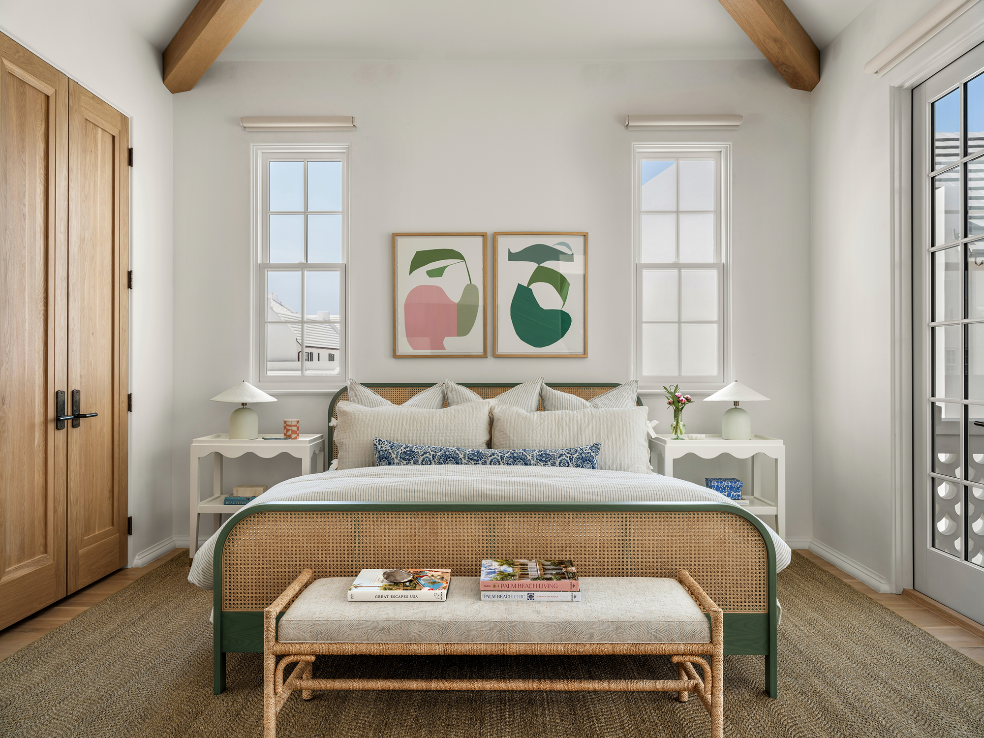 Bright bedroom with white walls, wooden ceiling beams, a bed with white and blue pillows, two white nightstands with lamps, two framed abstract art pieces above the bed, a woven bench at the foot of the bed with magazines, a woven rug, wooden closet 