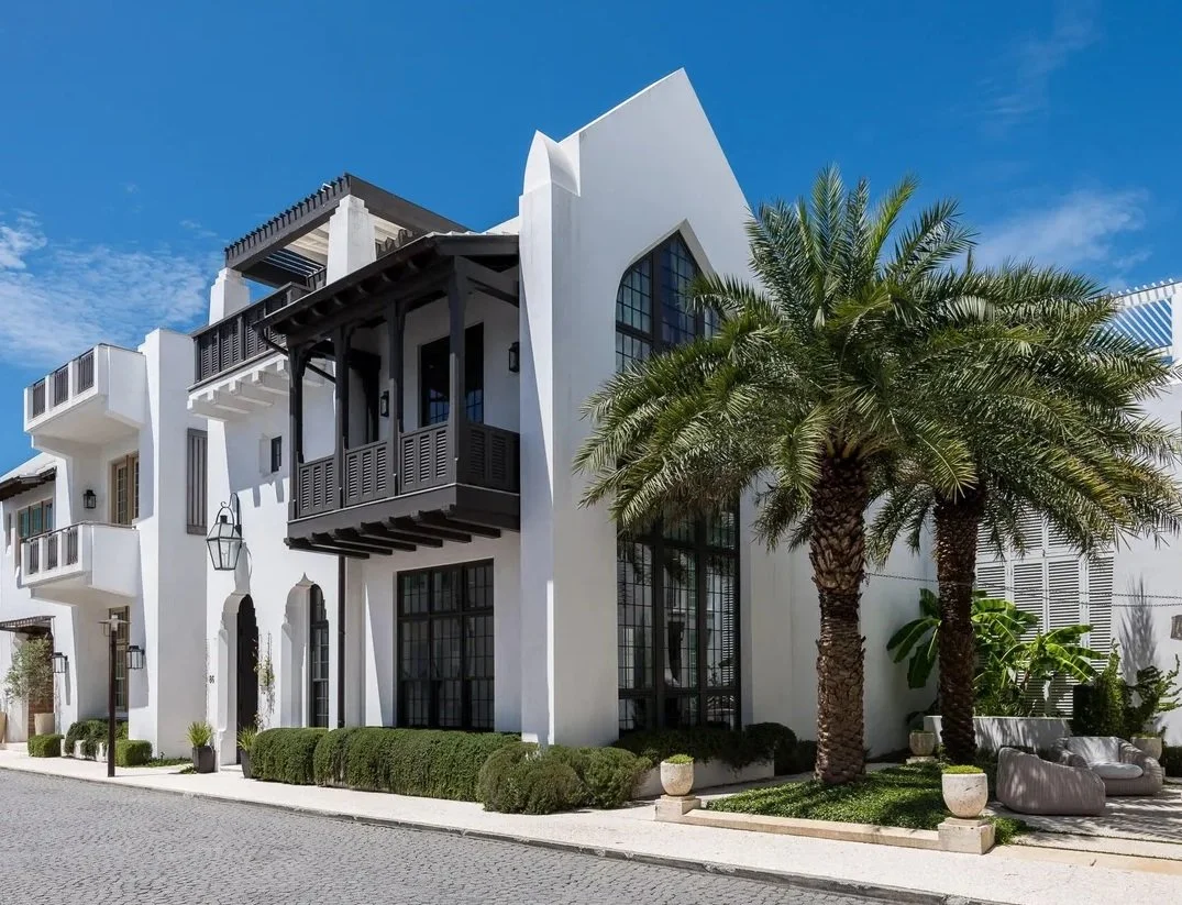 A white modern apartment building with black balconies and large windows, flanked by palm trees and outdoor seating, under a blue sky.