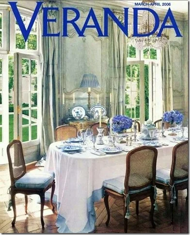 Cover of Veranda magazine featuring a formal dining room with a round table, white tablecloth, blue floral centerpiece, and elegant chairs, with large windows and light blue curtains in the background.