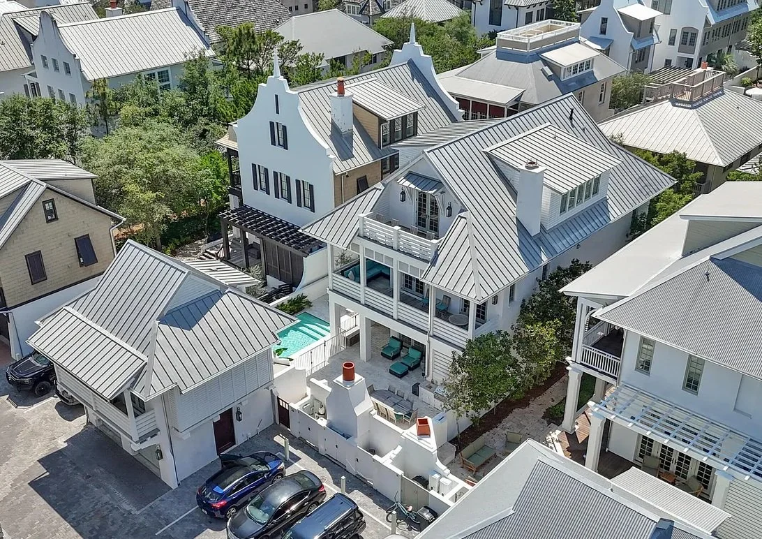Aerial view of white and gray residential houses with metal roofs, outdoor patios, and parking spaces, surrounded by trees and neighboring homes.
