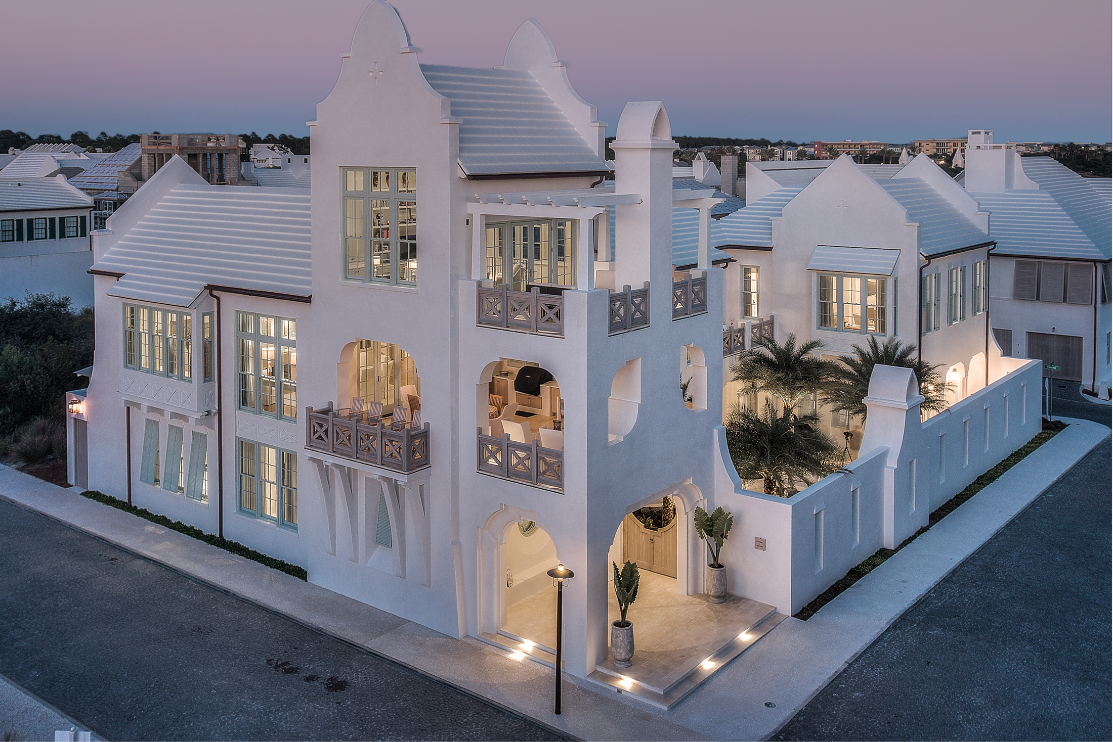 A large, white, modern house with multiple stories, arched doorways, and large windows, illuminated at dusk.