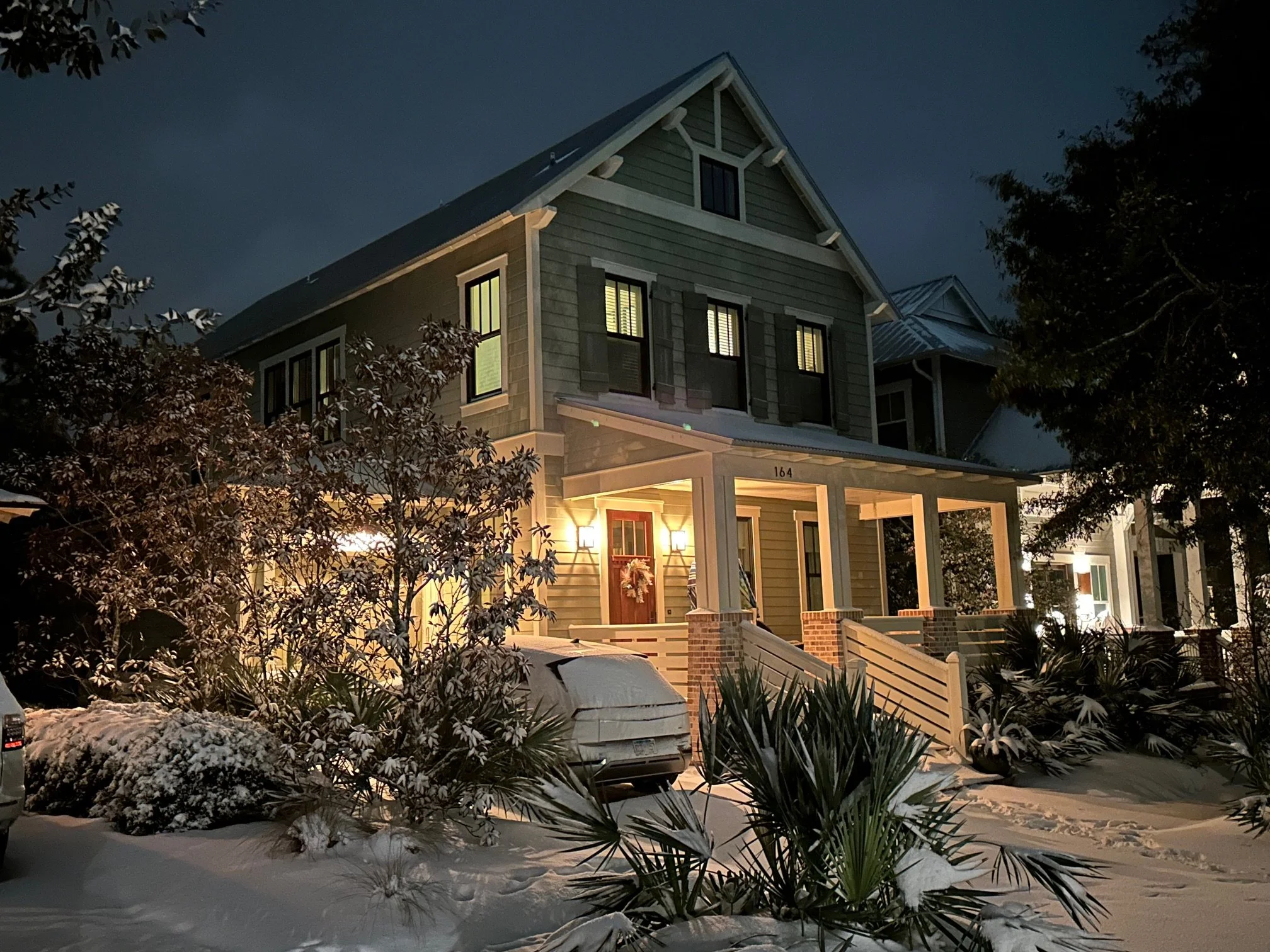 A two-story house illuminated at night with snow on the ground and surrounding plants, with trees and neighboring houses in the background.