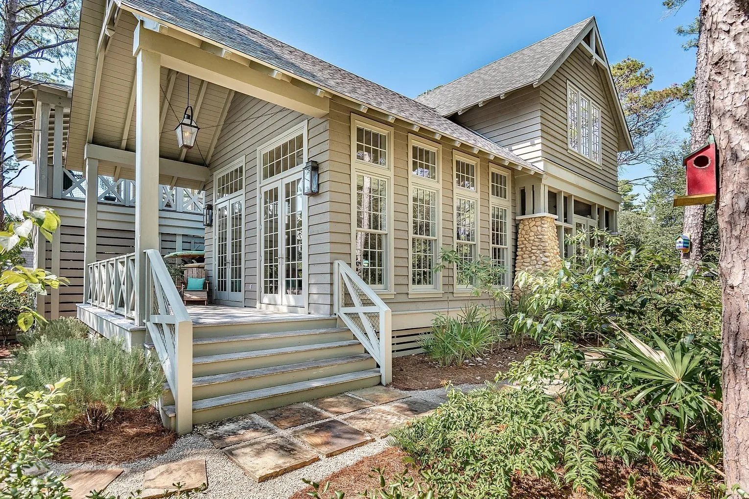 Two-story house with a wooden exterior, multiple large windows, staircase leading to a porch, surrounded by greenery, and a birdhouse attached to a tree.