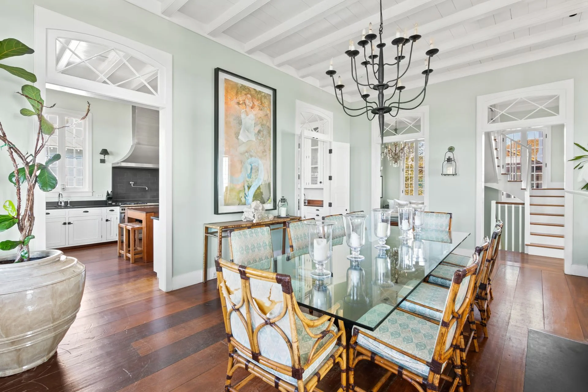 Bright dining room with a large glass table, upholstered chairs, a black chandelier, and a large framed artwork on pastel green walls. Visible kitchen with white cabinets and dark countertops, and a staircase in the background.