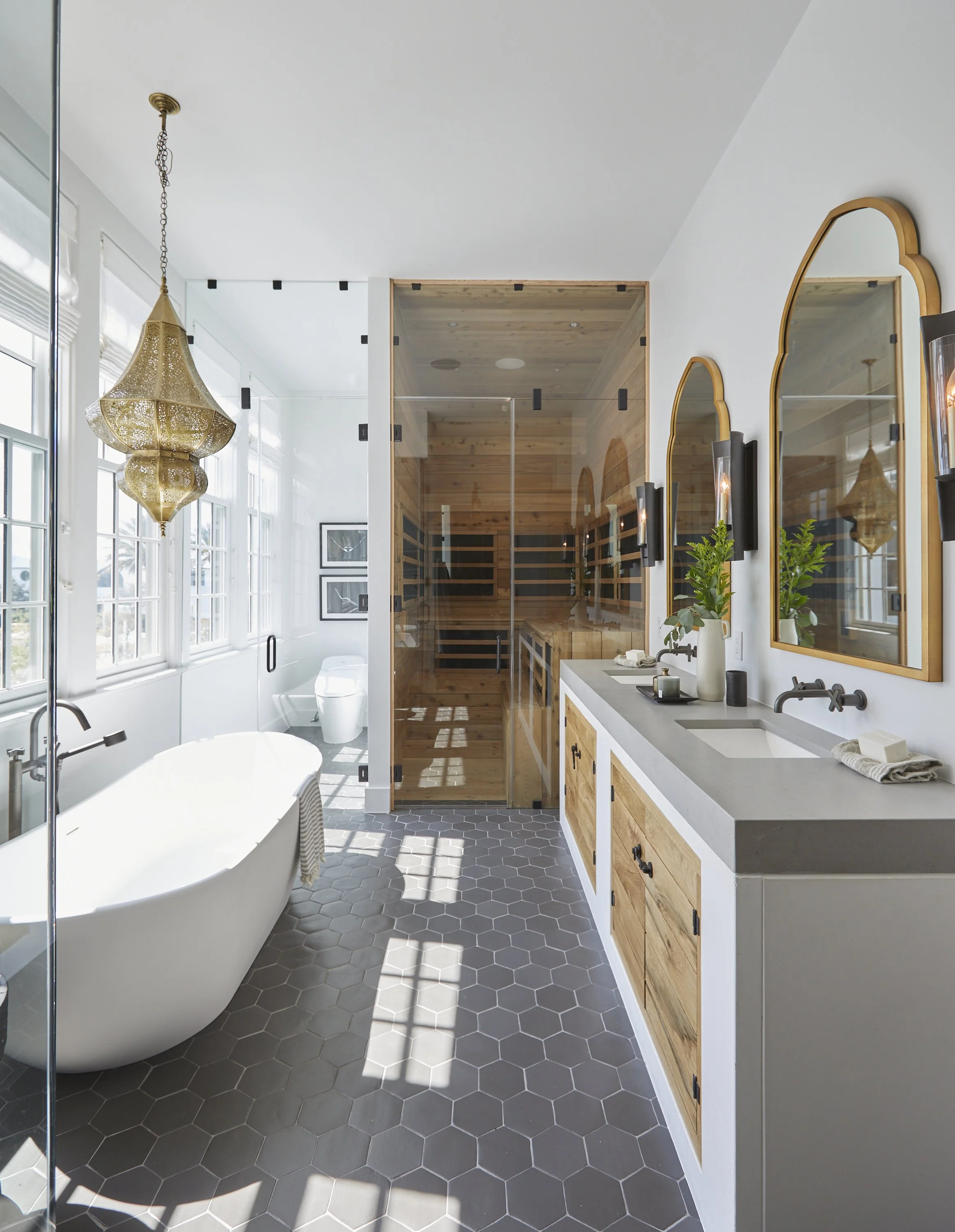 Modern bathroom with a white bathtub, wooden vanity with two mirrors, and a separate glass sauna in the background. Sunlight streams through the windows, creating shadows on the hexagonal gray tile floor. Decor includes a gold chandelier and green pl