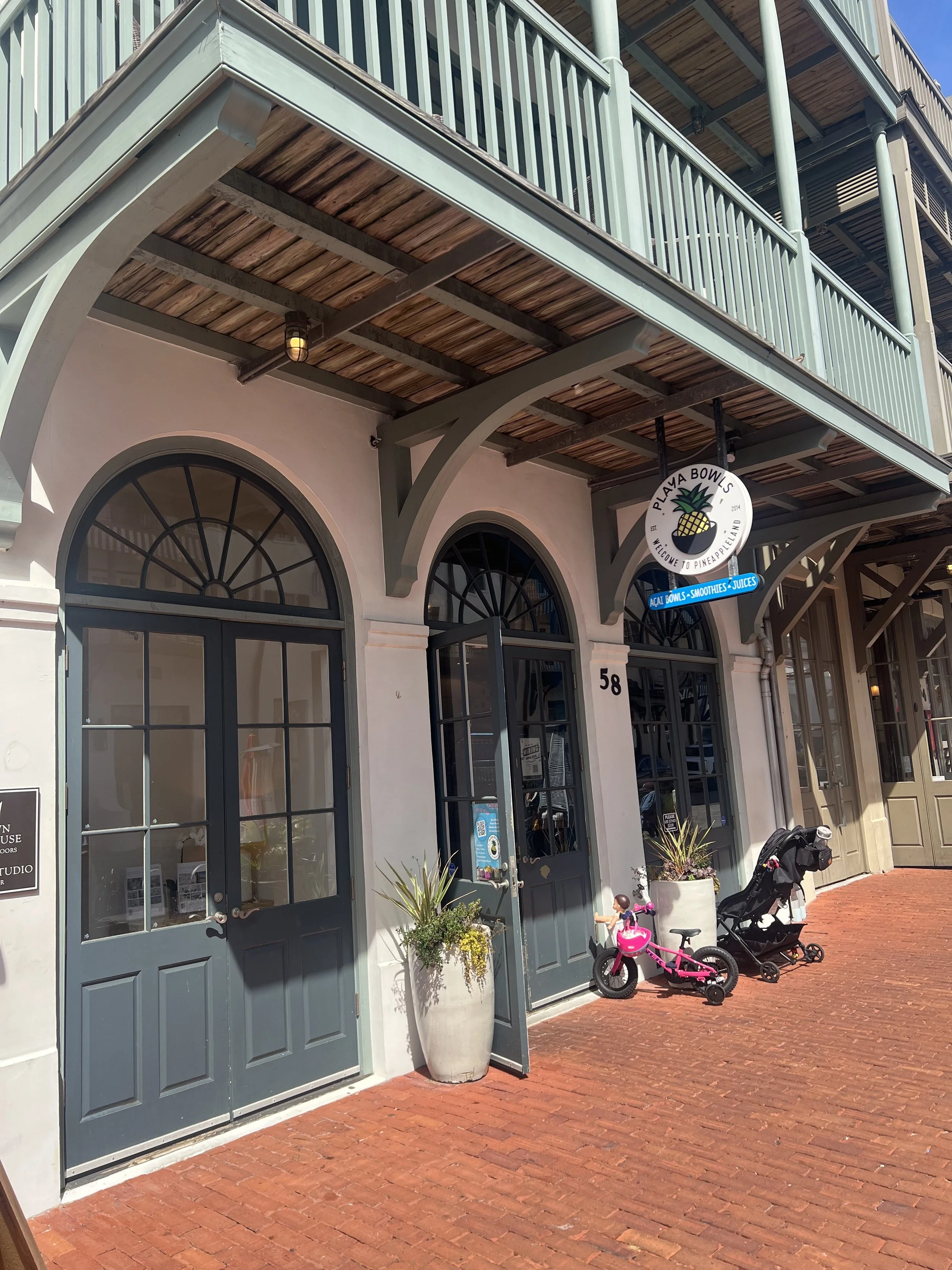 Exterior view of a building housing Playa Bowls with a sign showing it sells açaí bowls, smoothies, and juices. The building has arched windows and doors painted in dark blue, with potted plants outside. A pink children's bike and a black stroller ar