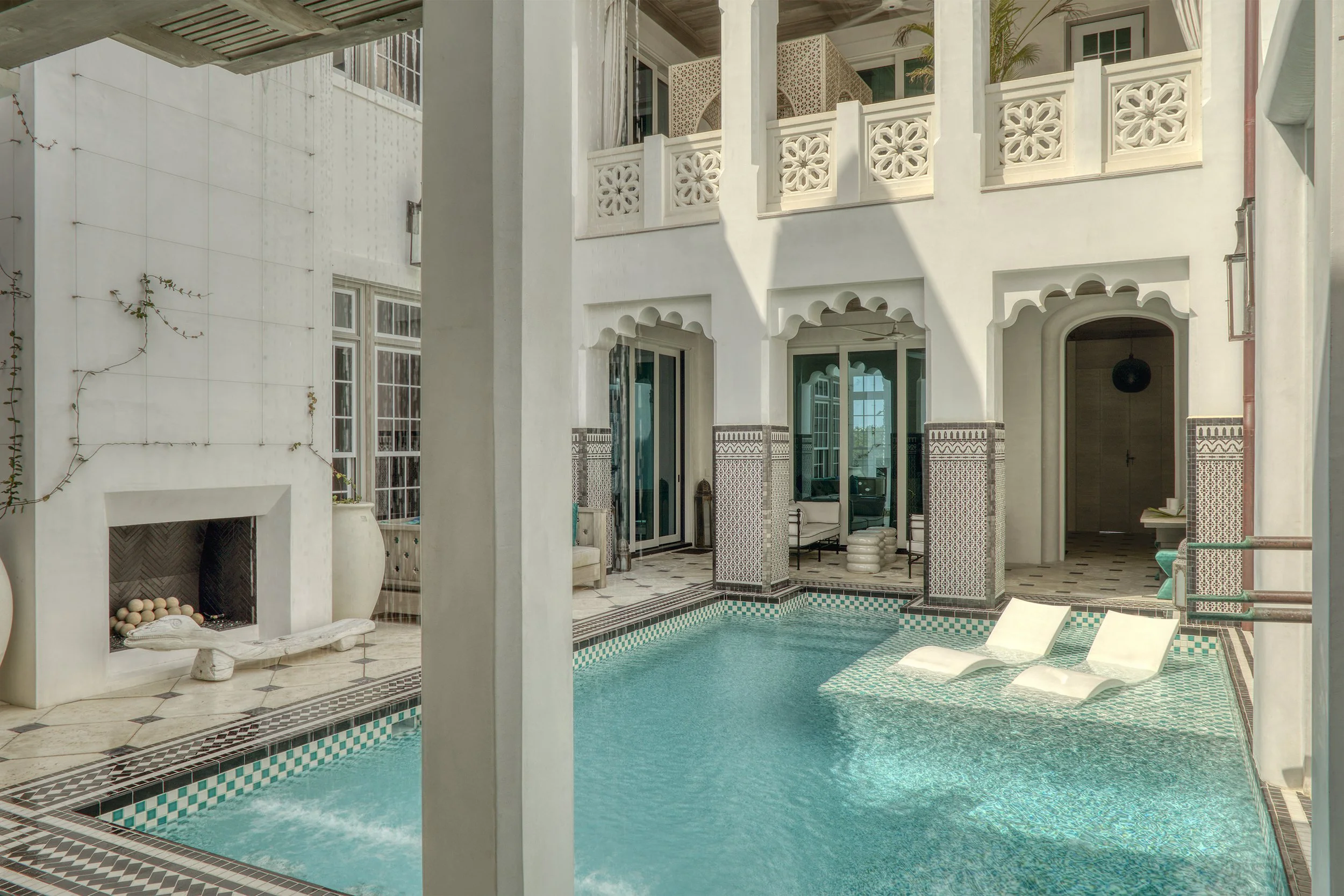 An outdoor pool area with two white lounge chairs partially submerged in a corner of the pool. The surrounding architecture features white walls, arched doorways, decorative tiles, and a small seating area with chairs and a table. There is a fireplace with decorative eggs on top and large potted plants.