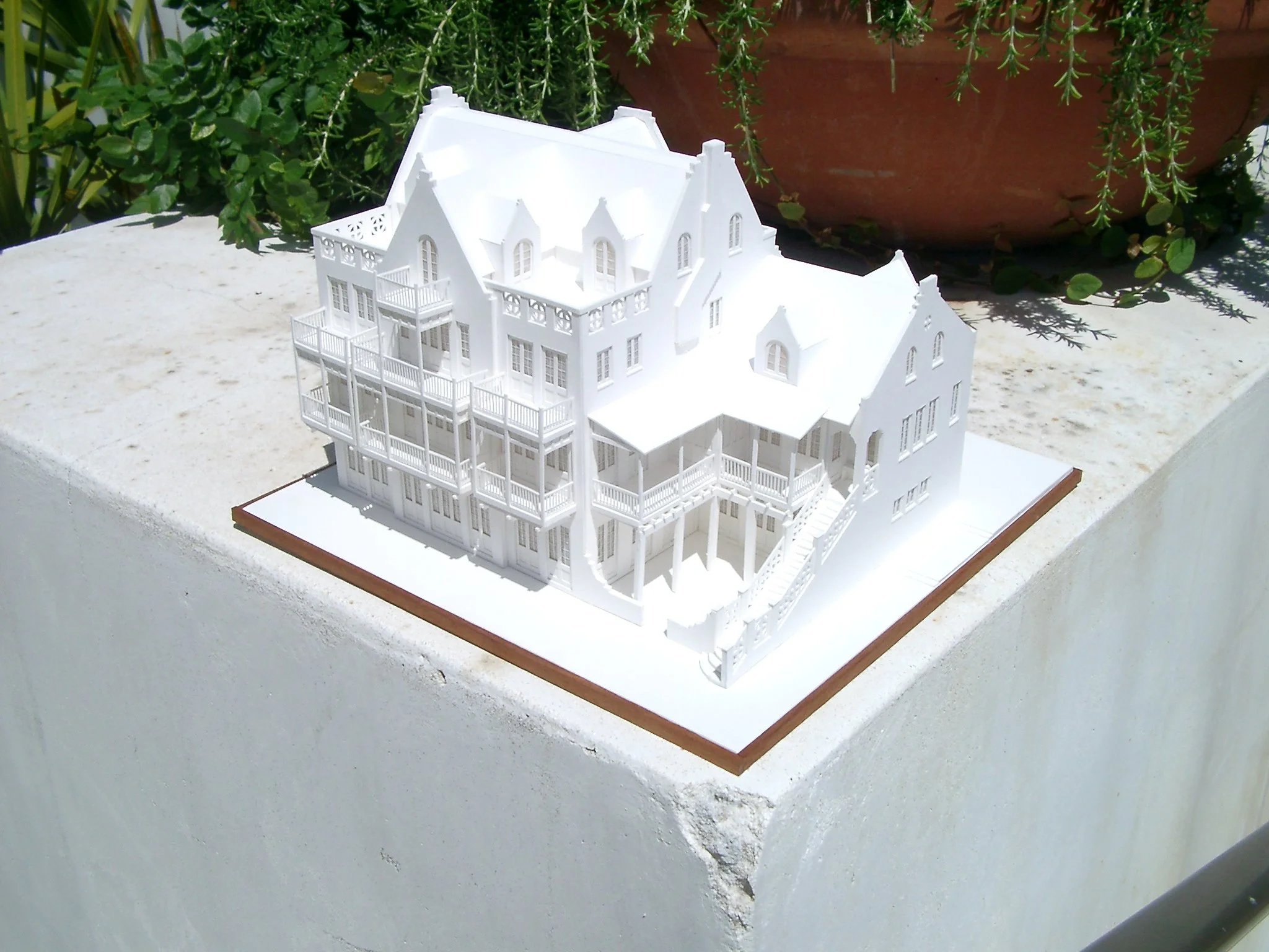 A white architectural model of a large, multi-story house with multiple balconies, windows, and a staircase, placed on a white surface outdoors, with potted plants in the background.