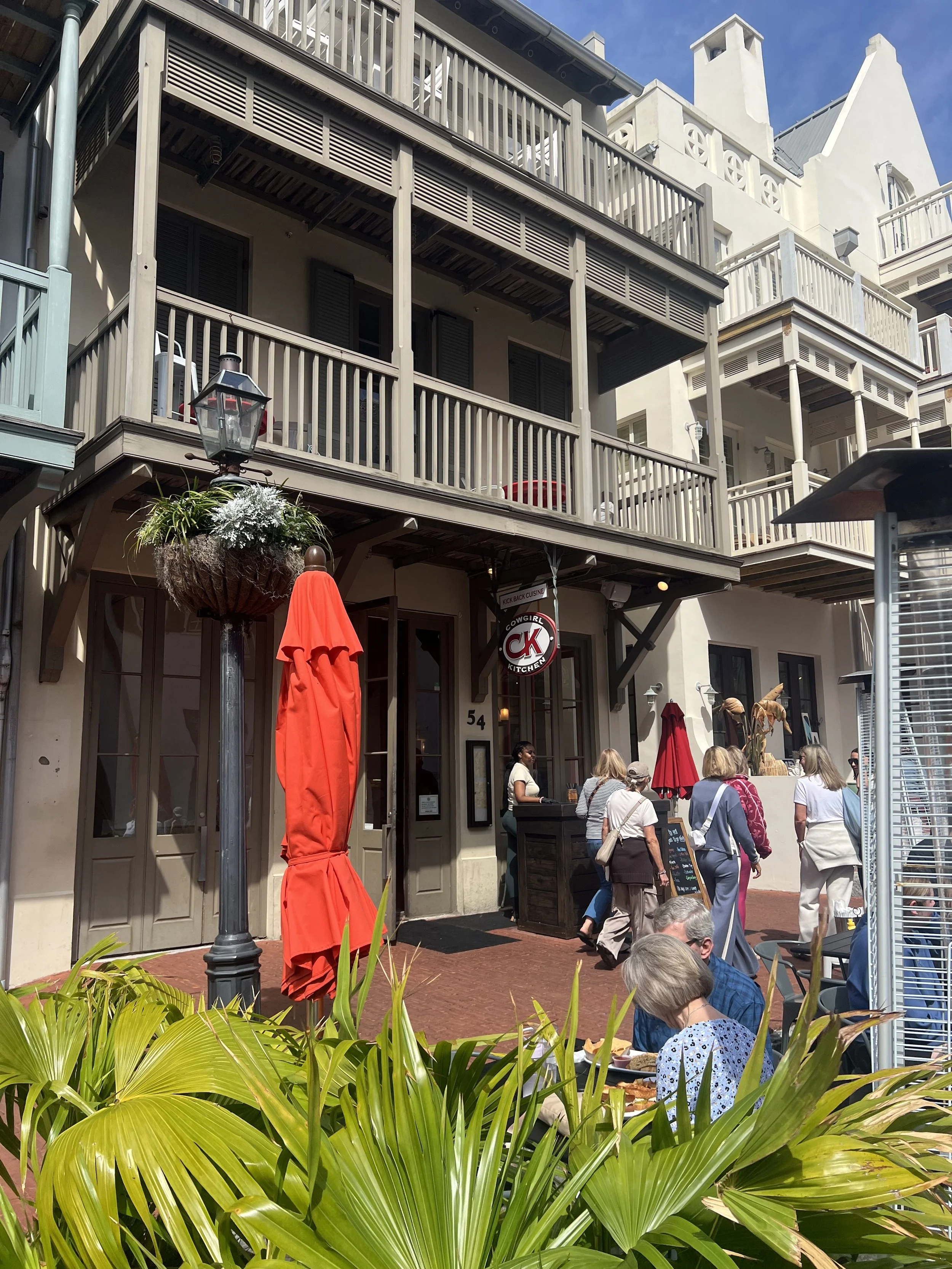 People dining outside a restaurant with a sign reading 'Cowgirl Kitchen,' in front of a multi-story building with balconies.
