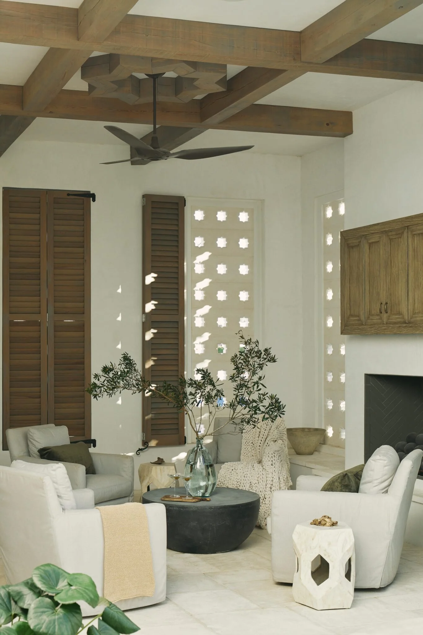 A living room with a mid-century modern style, featuring white armchairs, a dark round coffee table, a large glass vase with greenery, patterned pillows, a white side table with a decorative object, wooden window shutters, and a ceiling fan with wooden beams.
