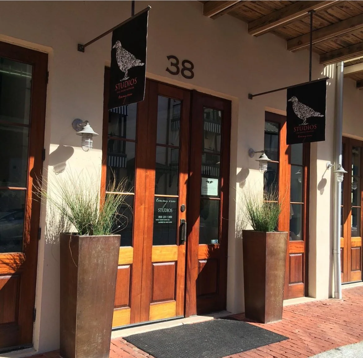 Front entrance of a building with wooden doors, two large planters with grass, and black hanging signs with a bird design and the word "STUDIOS".
