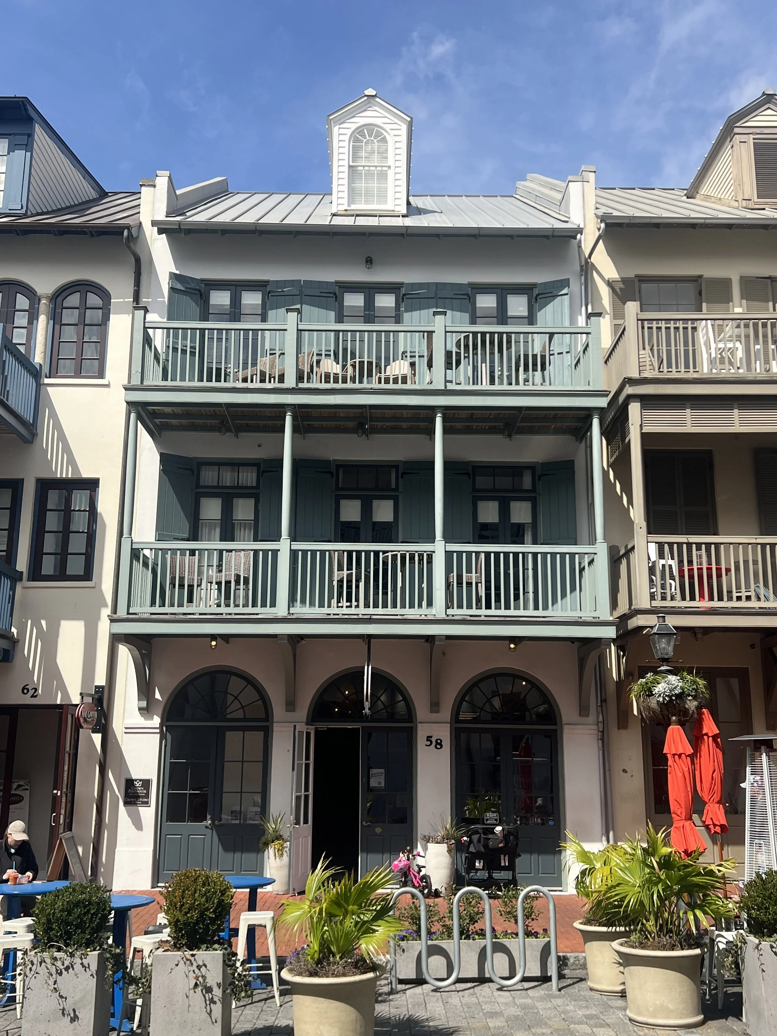 A three-story building with light blue and cream exterior, featuring balconies on the second and third floors, large arched windows on the ground floor, and outdoor seating with potted plants and red umbrellas.