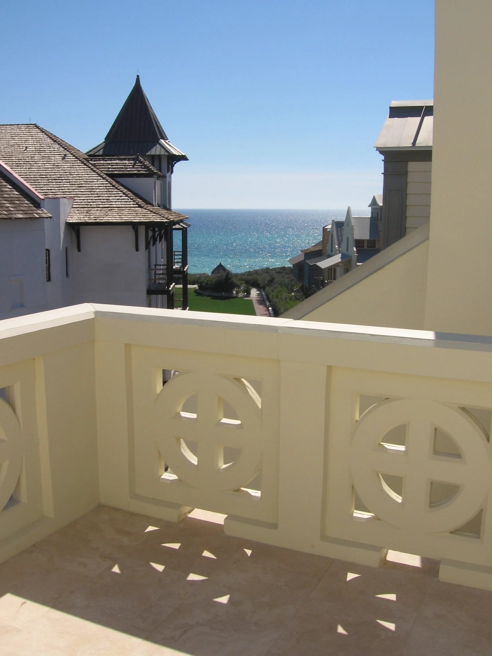 View from a balcony overlooking a neighborhood with houses, some with pointed rooftops and a tower, leading to the ocean in the distance under a clear blue sky.