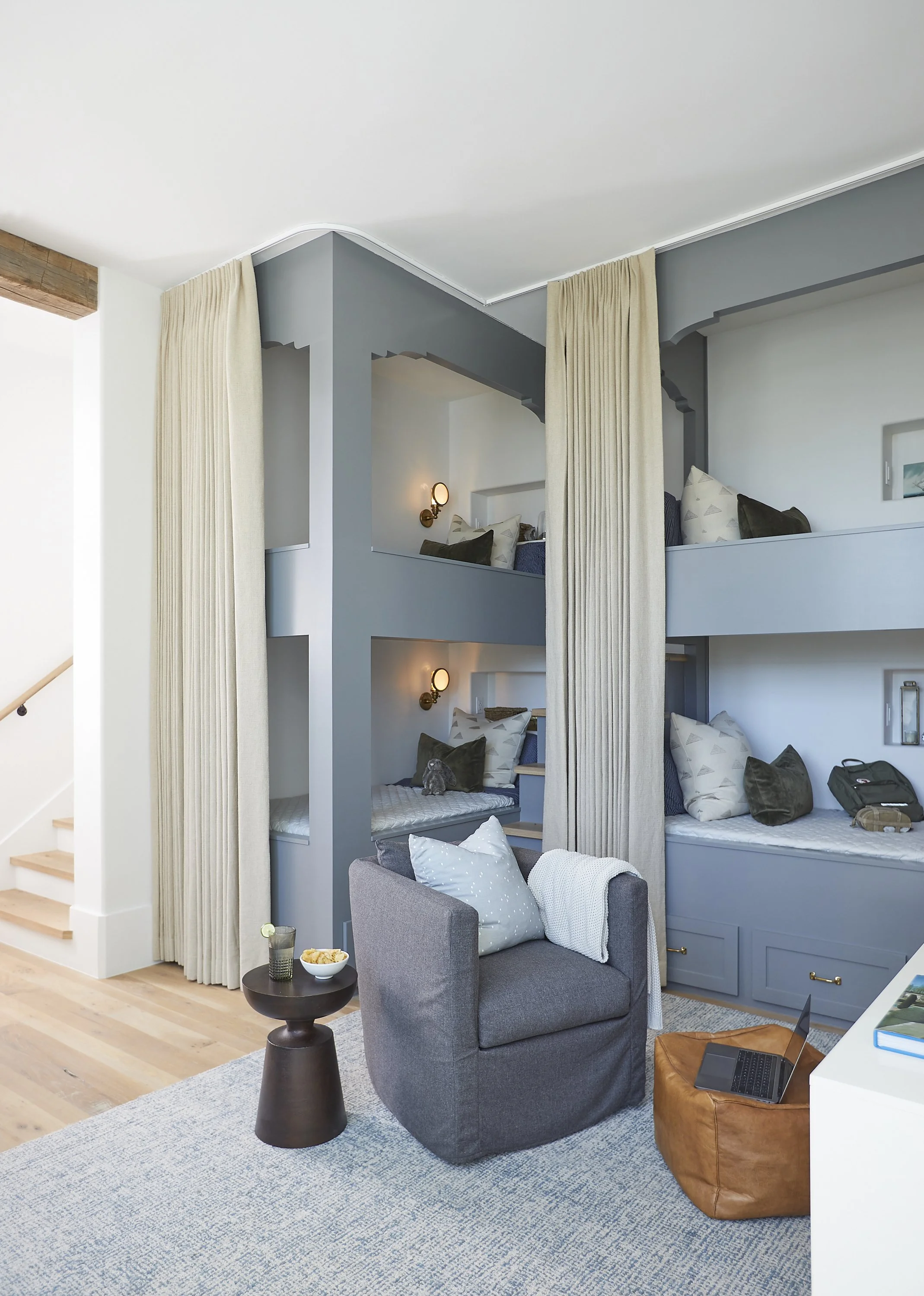 Interior of a hotel room with two bunk beds separated by curtains, gray walls, and a sitting area with a gray armchair, a small side table, and a leather ottoman with a laptop on it.