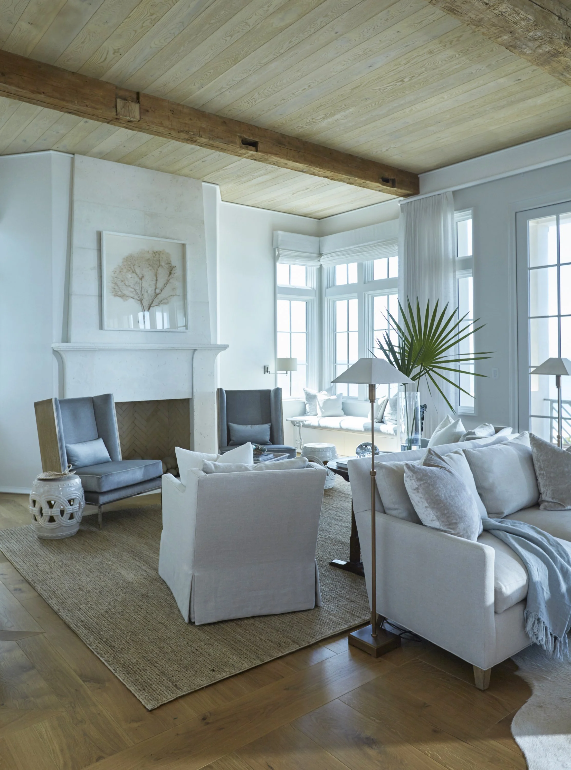Cozy living room with white walls, large windows, a fireplace, light gray armchairs, a white sofa, a large green potted plant, table lamps, a beige area rug, and wooden ceiling beams.
