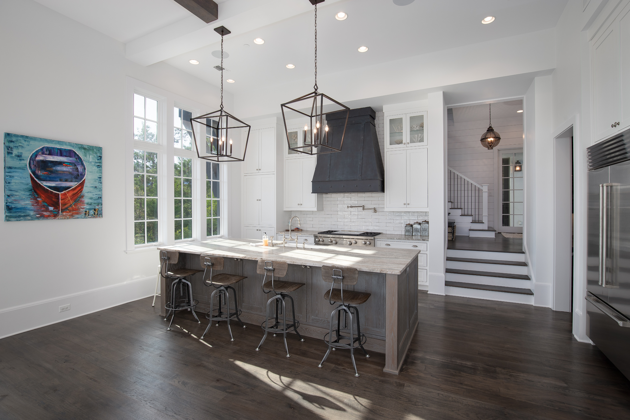 Modern kitchen with white cabinets, dark wood flooring, and a large island with seating. Pendant lights hang above the island. There are large windows letting in natural light. A painting of a boat is on the wall, and stairs are visible in the backgr