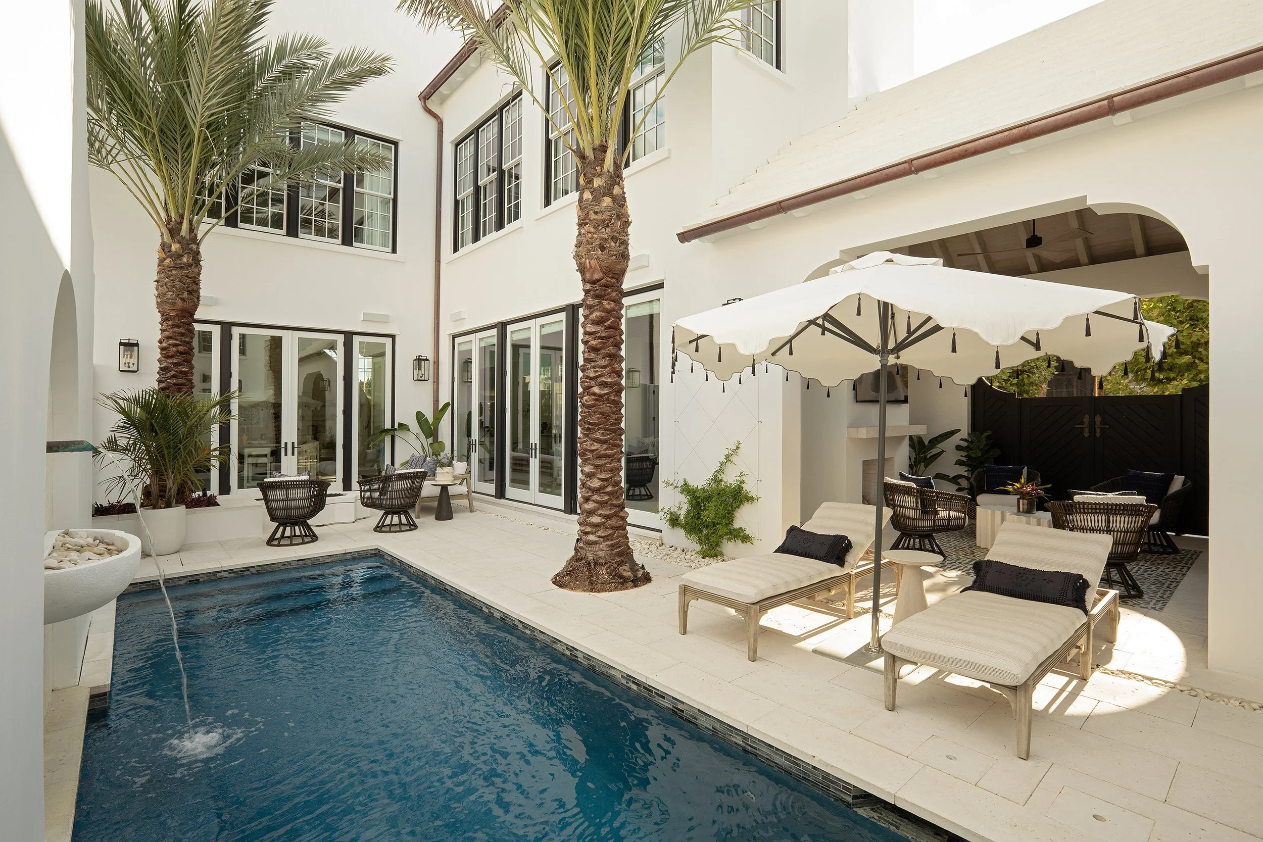 A luxurious backyard with a swimming pool, white patio furniture, palm trees, and an umbrella providing shade. The space is surrounded by white building walls with large windows and glass doors.