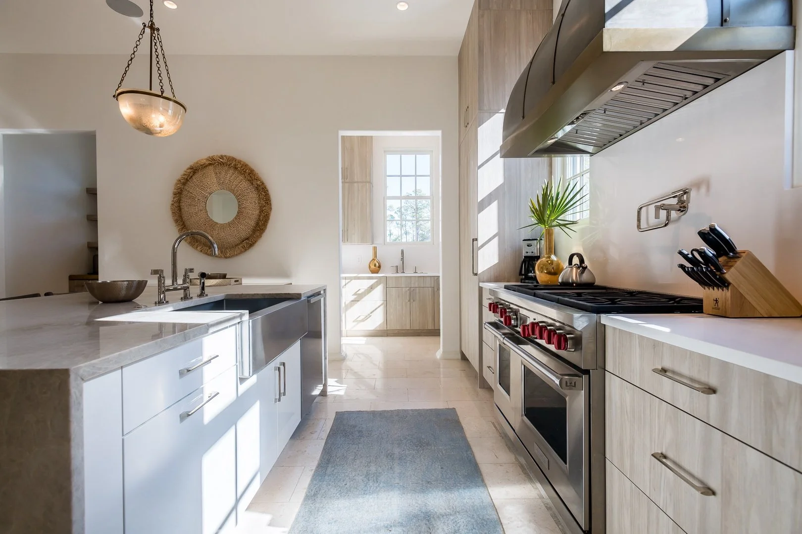 Bright kitchen featuring a large island with a marble countertop, a stainless steel sink, and modern cabinets. To the right, a stainless steel stove with red knobs, a wooden knife block, and a potted plant in a gold vase. Natural light streams through a window into a smaller adjacent room with more cabinetry and decor.