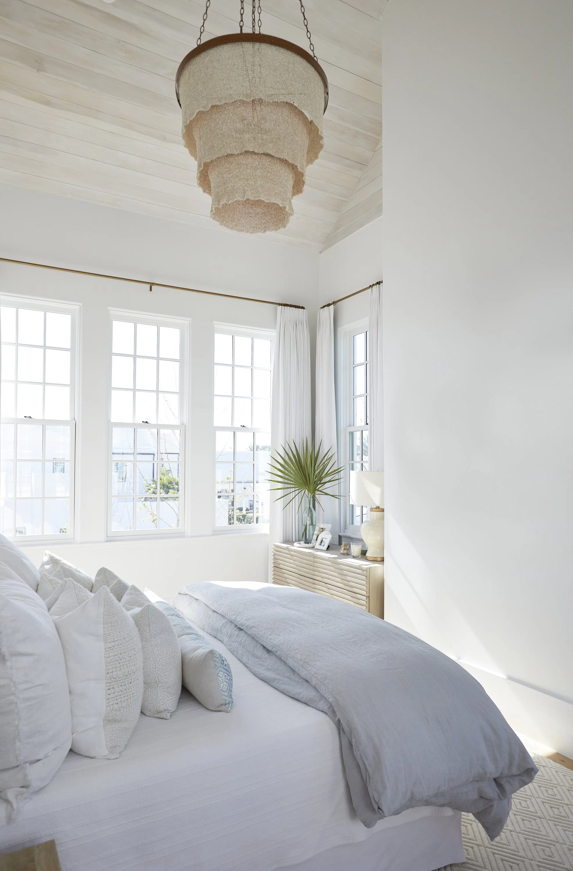 Bright bedroom with white walls, multiple windows with white curtains, a white bed with multiple pillows, beige bedroom dresser with a large palm leaf in a glass vase, a white table lamp, and a hanging beige chandelier.
