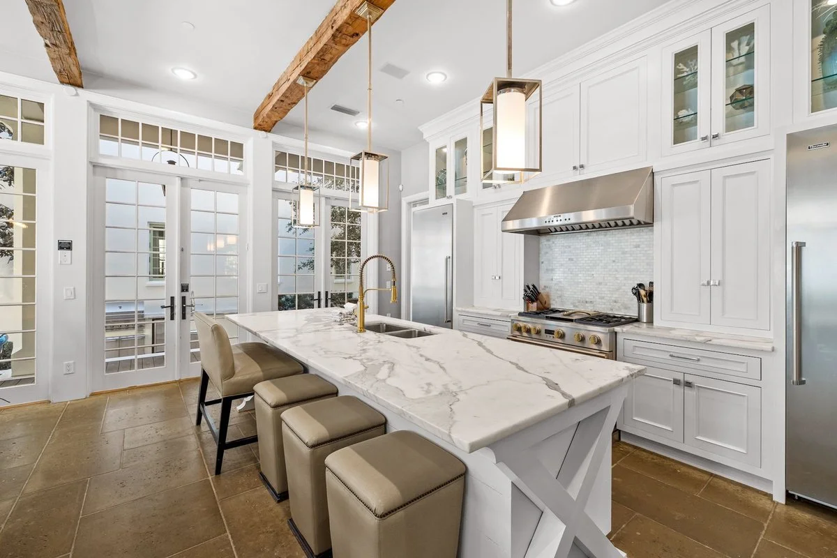 Modern white kitchen with marble island, stainless steel appliances, and glass-paneled cabinets, with wooden beams on the ceiling and large glass doors.