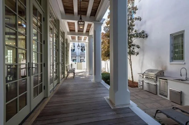 Covered patio with wooden flooring, glass-paneled doors, outdoor grill, sink, and potted trees.