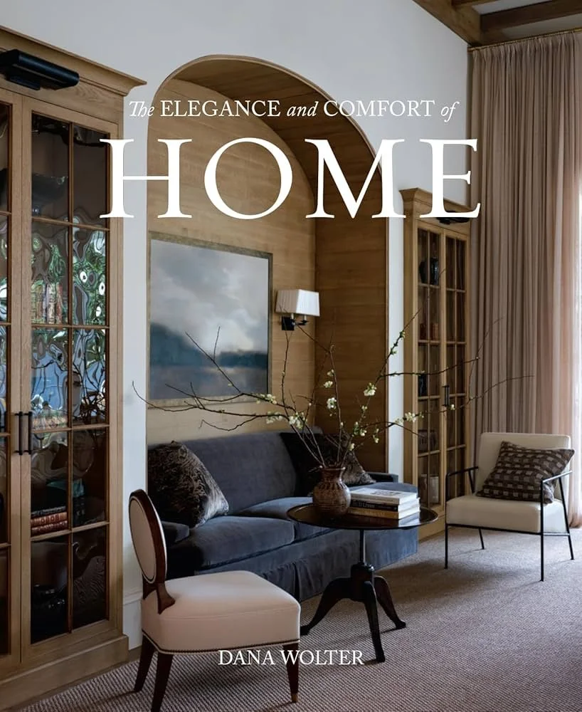 Cover of a home interior book featuring a cozy living room with a dark sofa, two white chairs, a wooden cabinet, and a table with a vase of branches and books. Text on the cover reads, 'The ELEGANCE and COMFORT of HOME' by Dana Wolter.