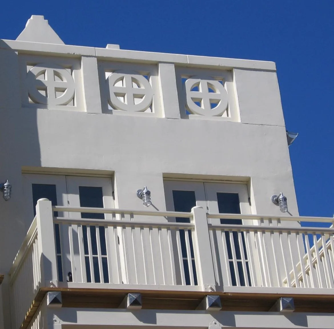 A white building with decorative circular window grills and a balcony with a white railing, against a clear blue sky.