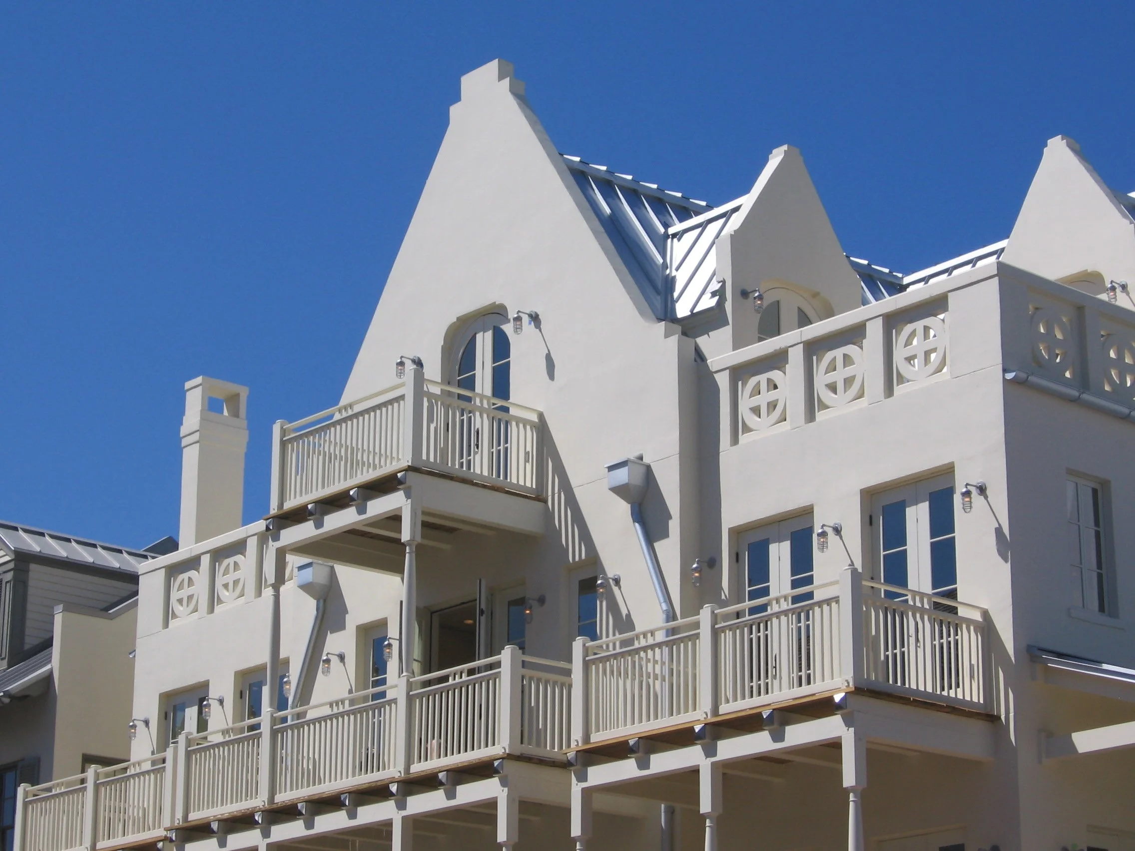 Close-up of a white multi-story building with balconies, arched windows, and a metal roof against a blue sky.