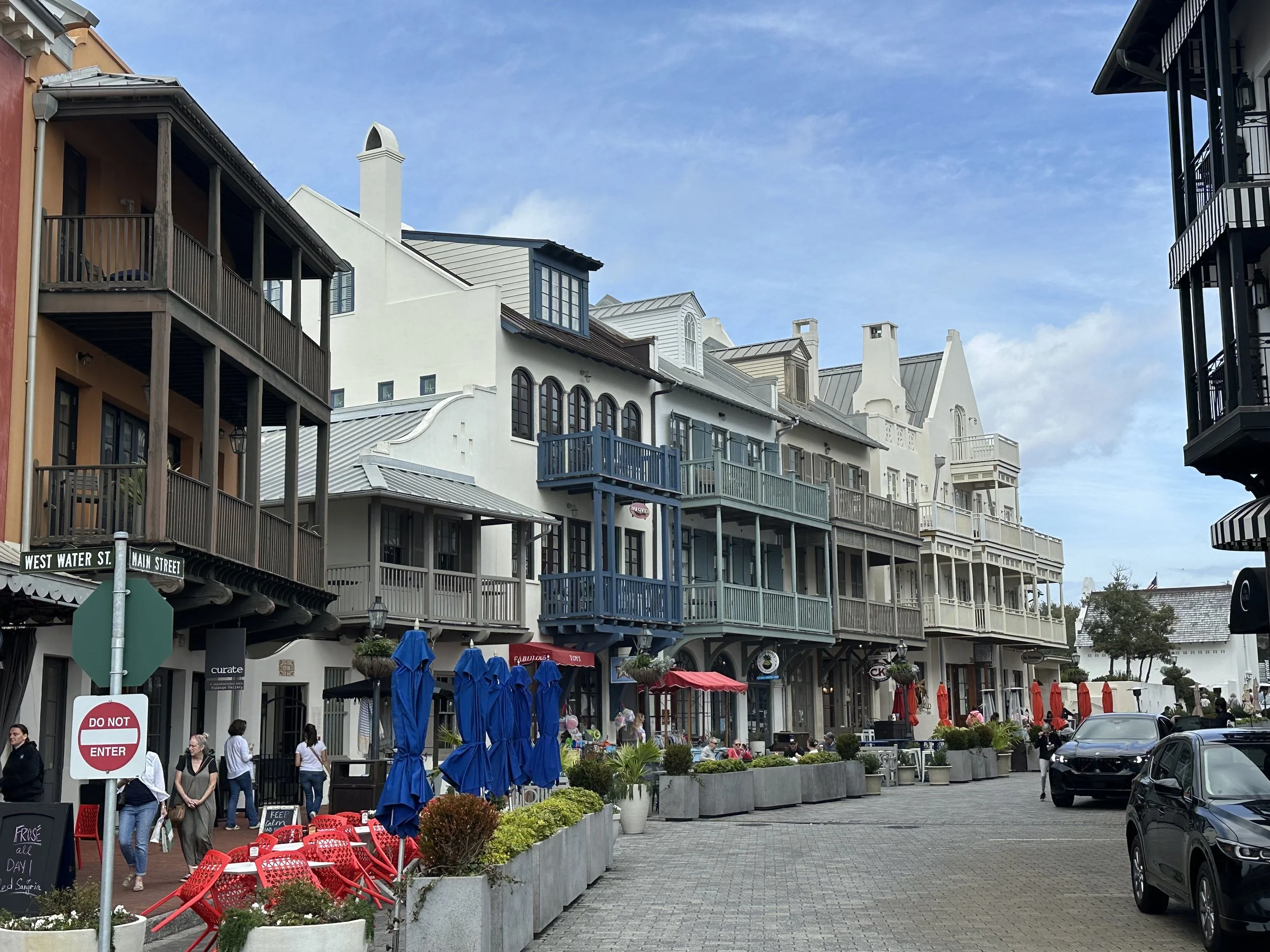 A street view of multi-story buildings with balconies, shops, and cafes, with people walking, outdoor seating with umbrellas, parked cars, and street signs indicating 'West Water St.' and 'Main Street.'