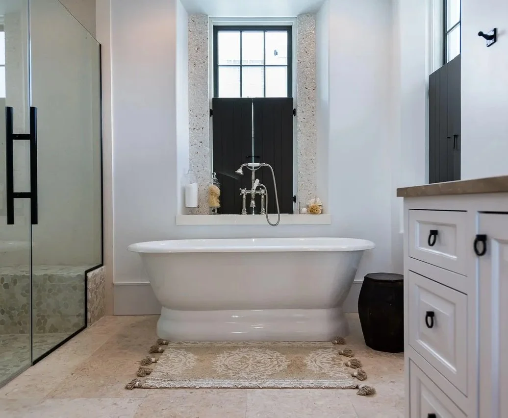 Bathroom with a clawfoot bathtub in front of a window, a glass-enclosed shower on the left, a white vanity on the right, and a small dark stool beside the tub.