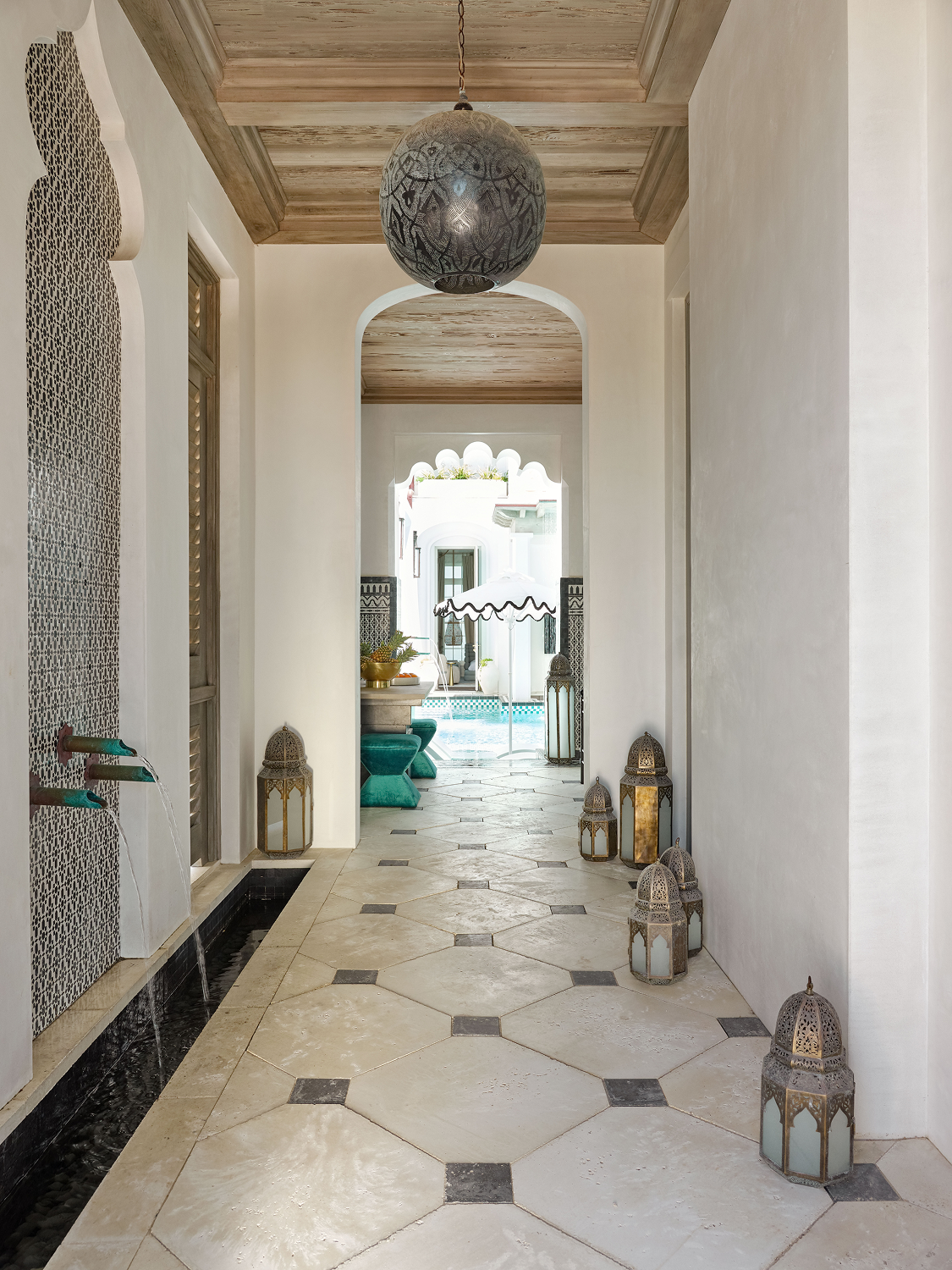Indoor corridor with white walls, tiled floor, hanging decorative lanterns, and view of outdoor pool and seating area with umbrella.