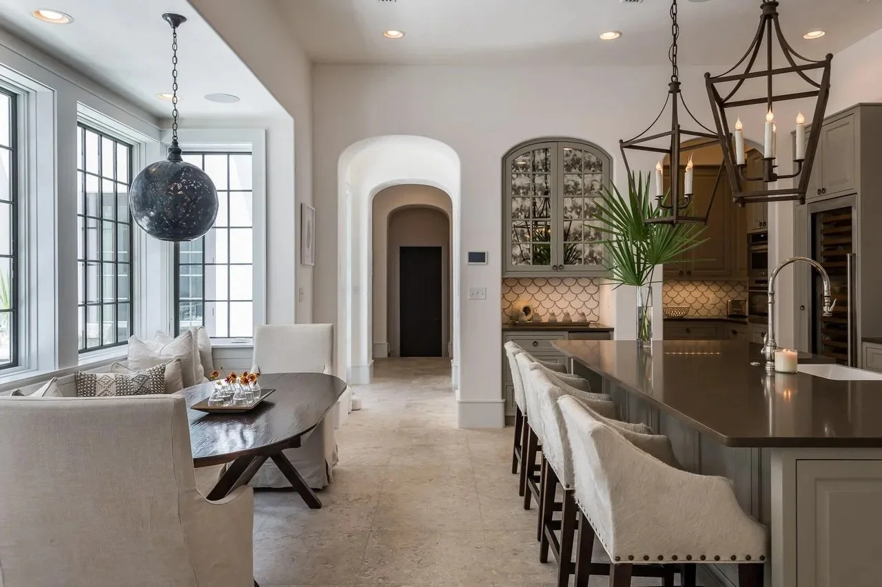 Modern kitchen and dining area with large windows, a dark pendant light, a wooden dining table with beige chairs, and a kitchen island with bar stools, pendant lighting, and a large green plant.