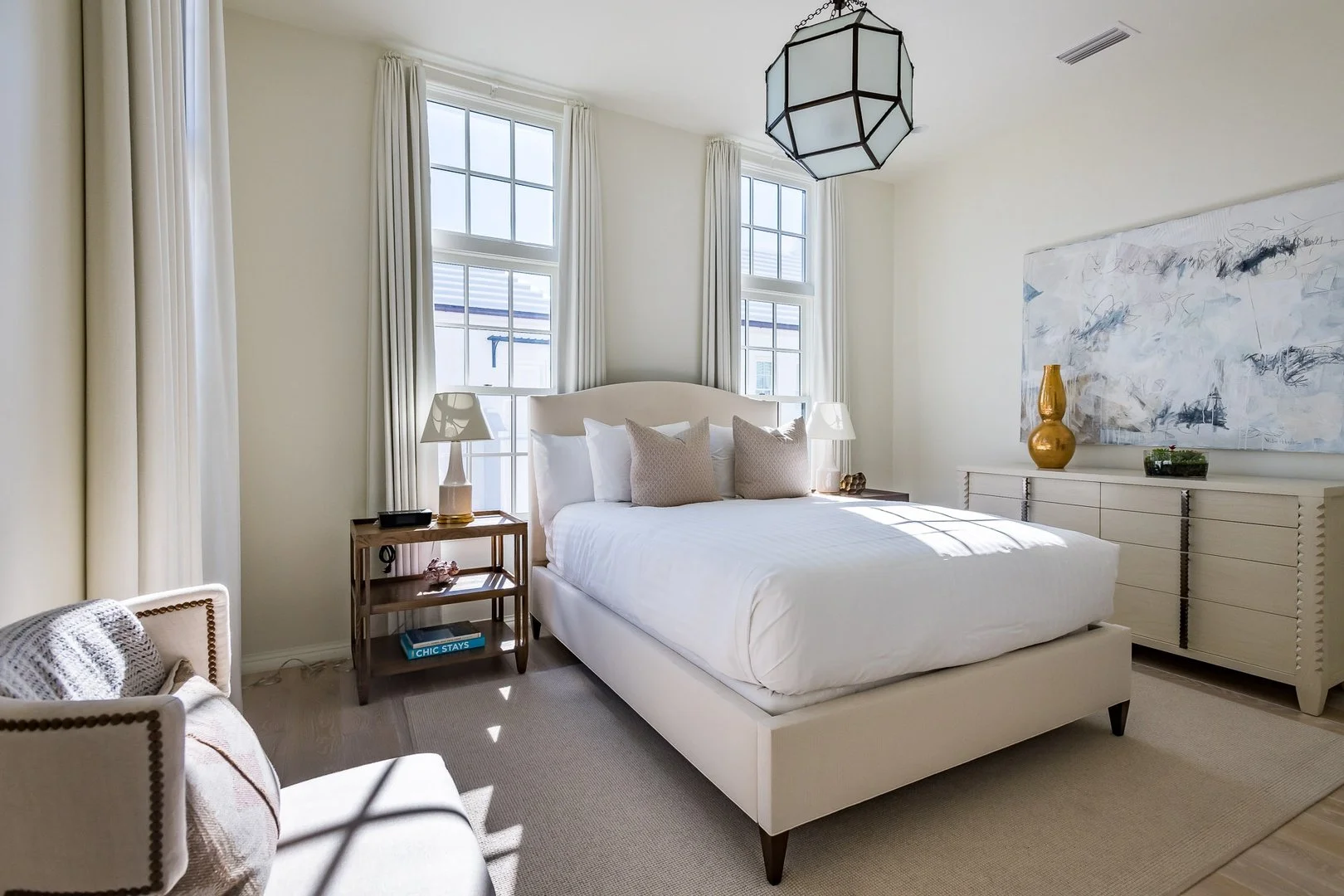 Bright, elegant bedroom with white bed, side tables, lamps, large windows with curtains, and modern decor