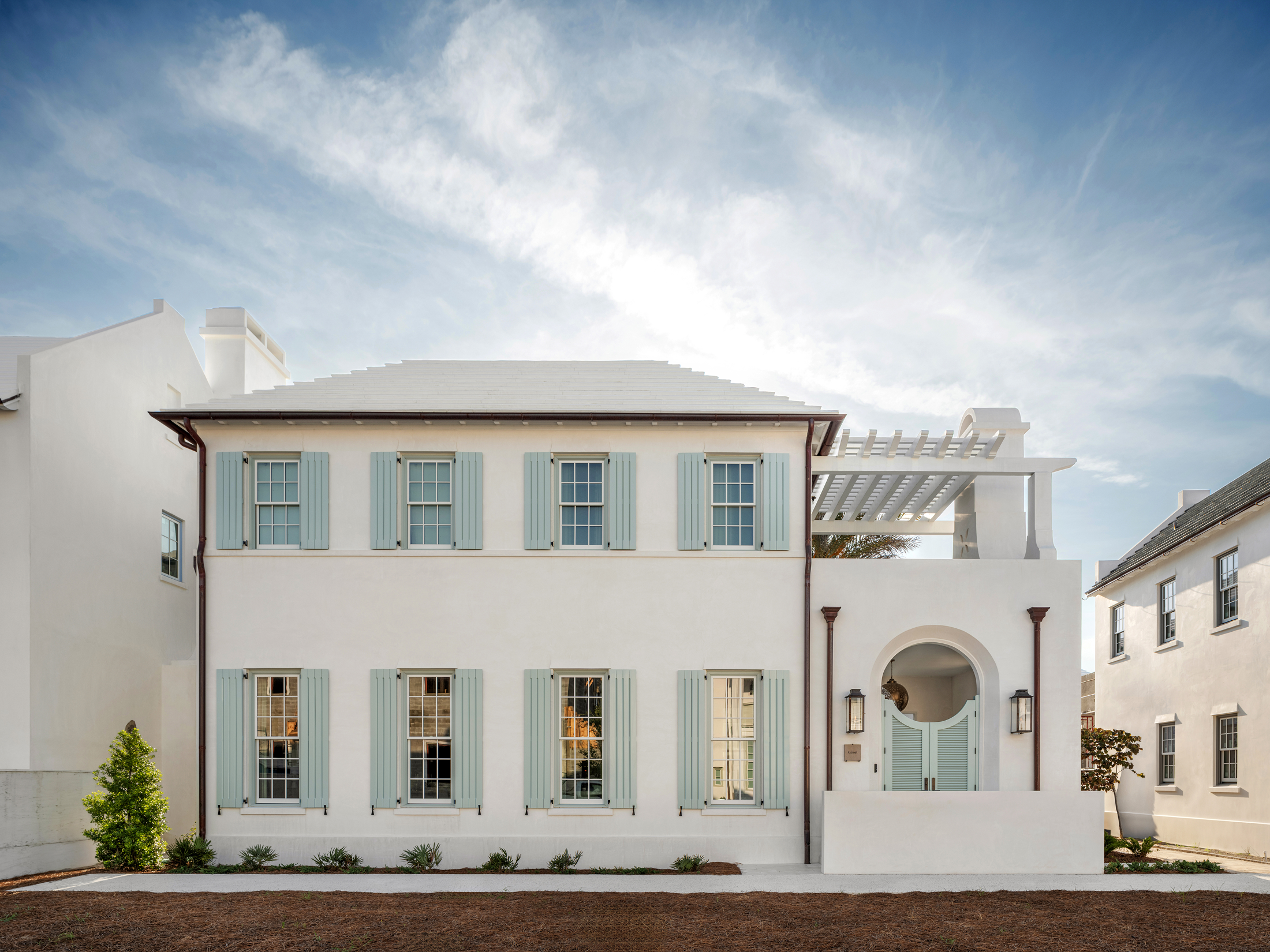 White two-story house with light blue shutters, arched doorway, and a pergola on the roof, set against a partly cloudy sky.