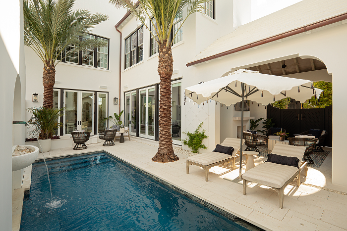 A luxurious backyard with a swimming pool, palm trees, lounge chairs with cushions and an umbrella, and outdoor seating area with tables and chairs in a modern house.