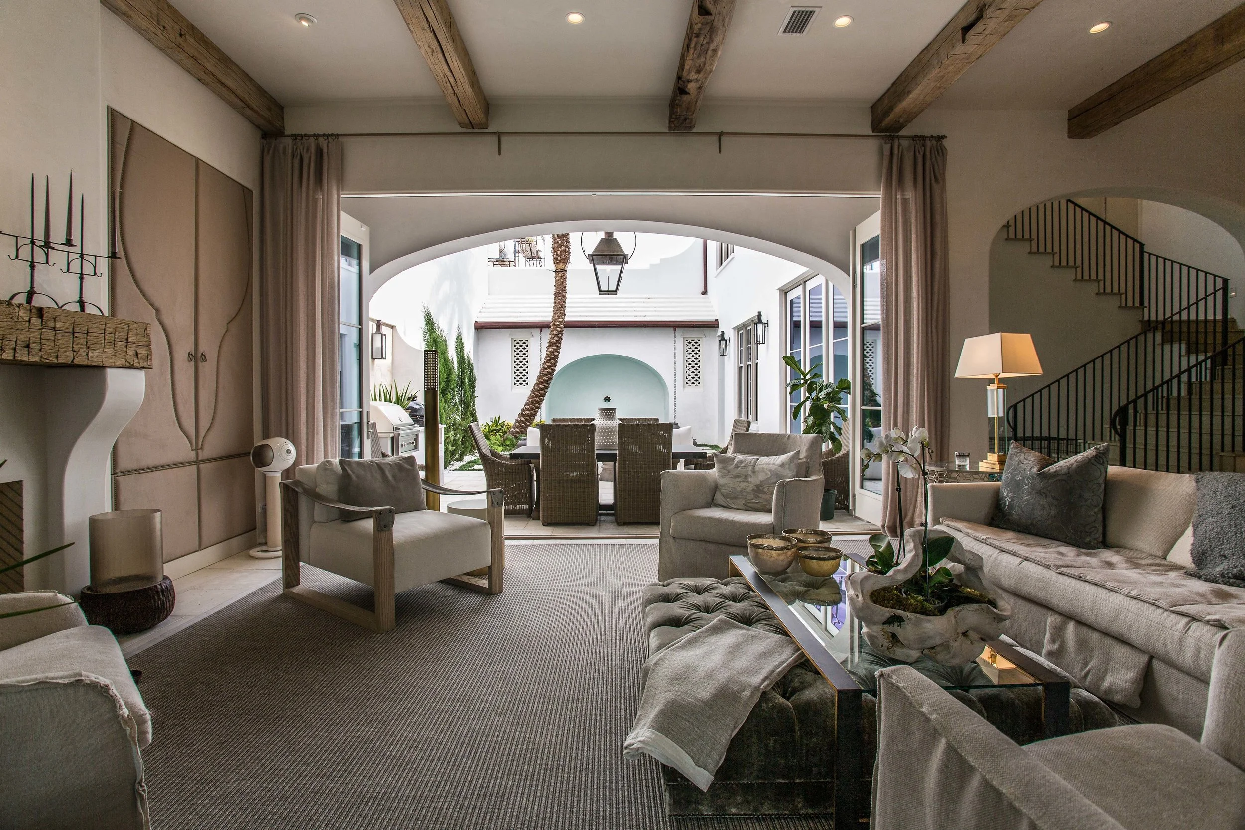 Living room with beige sofas and armchair, coffee table, and sliding glass doors opening to outdoor patio with dining table and chairs, palm tree, and decorative architecture.