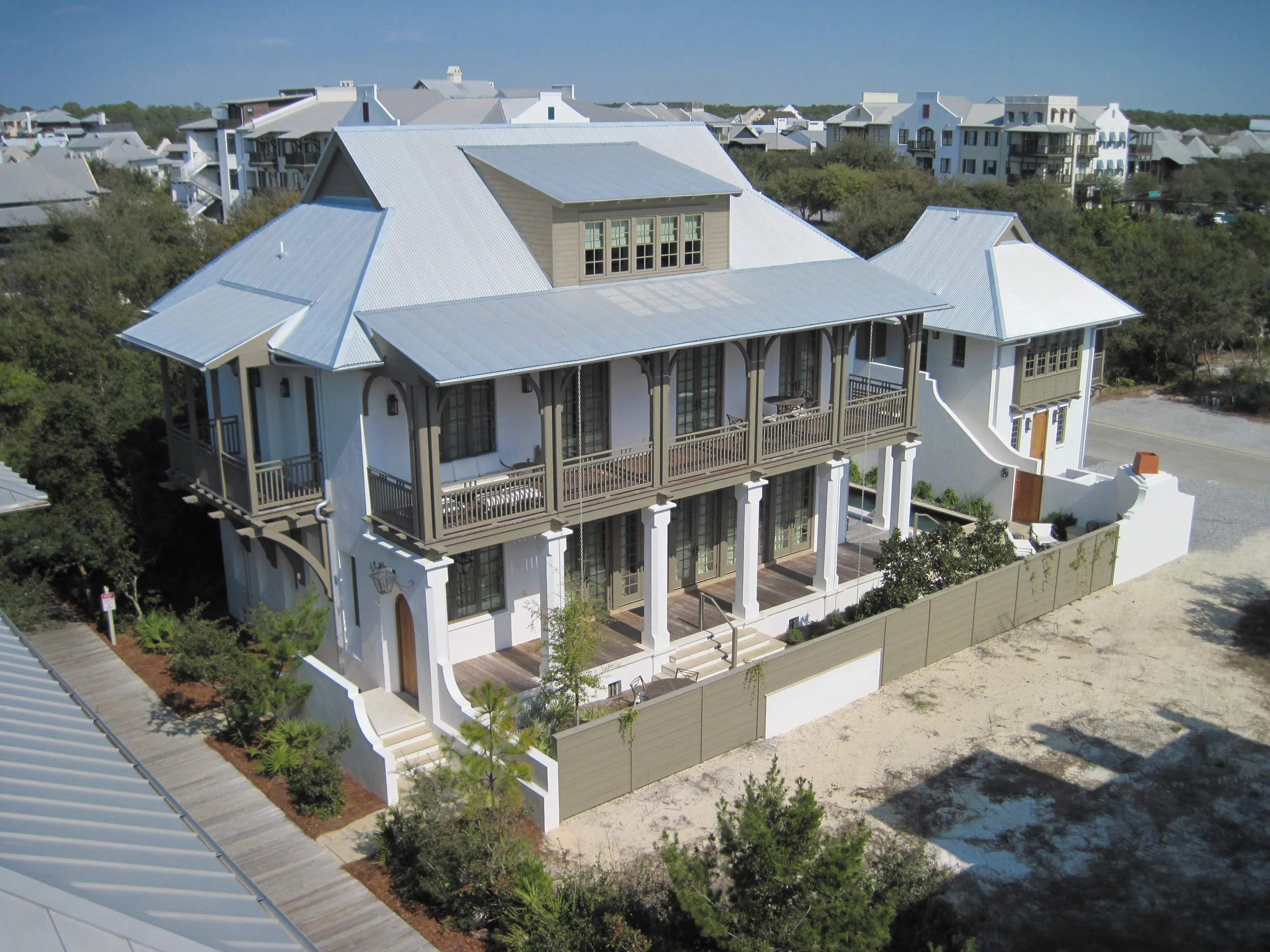 A large multi-story house with white walls, gray metal roofs, and a prominent balcony with columns and railings. Surrounding the house is a garden with trees and shrubs, and a wooden fence. Other similar houses are visible in the background.