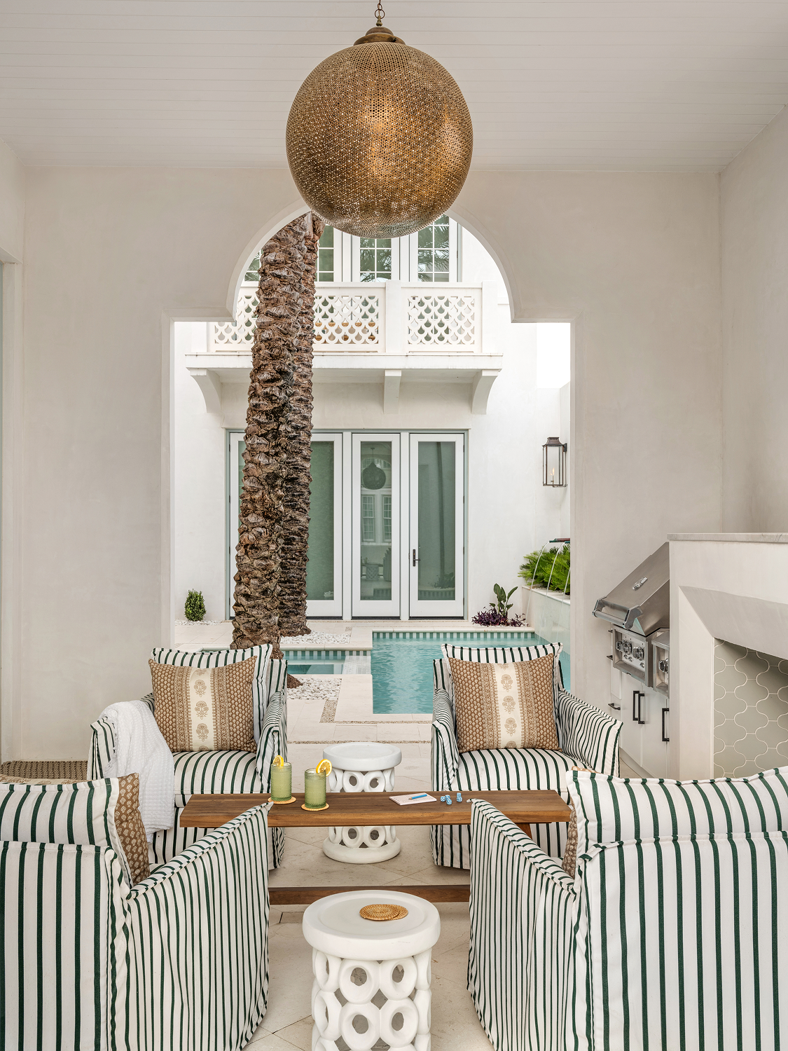 Indoor patio with striped armchairs, a wooden coffee table, small white side tables, a large hanging golden light fixture, a palm tree, a swimming pool, and glass doors leading outside.