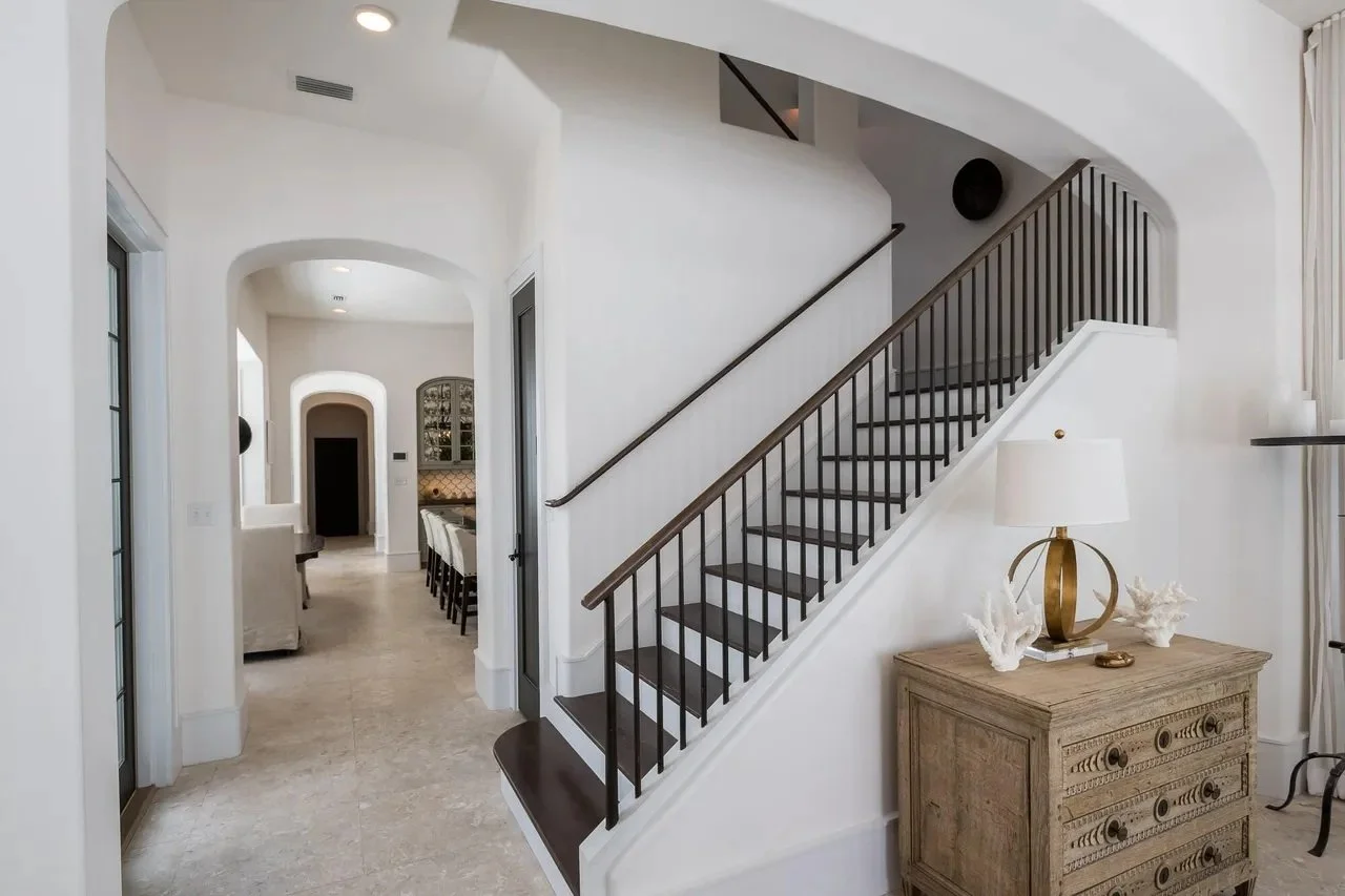A modern interior with a staircase having dark wooden steps and black railings next to a wooden dresser with a brass and white lamp and decorative coral pieces on top. The space is bright with white walls and a large window with sheer curtains on the