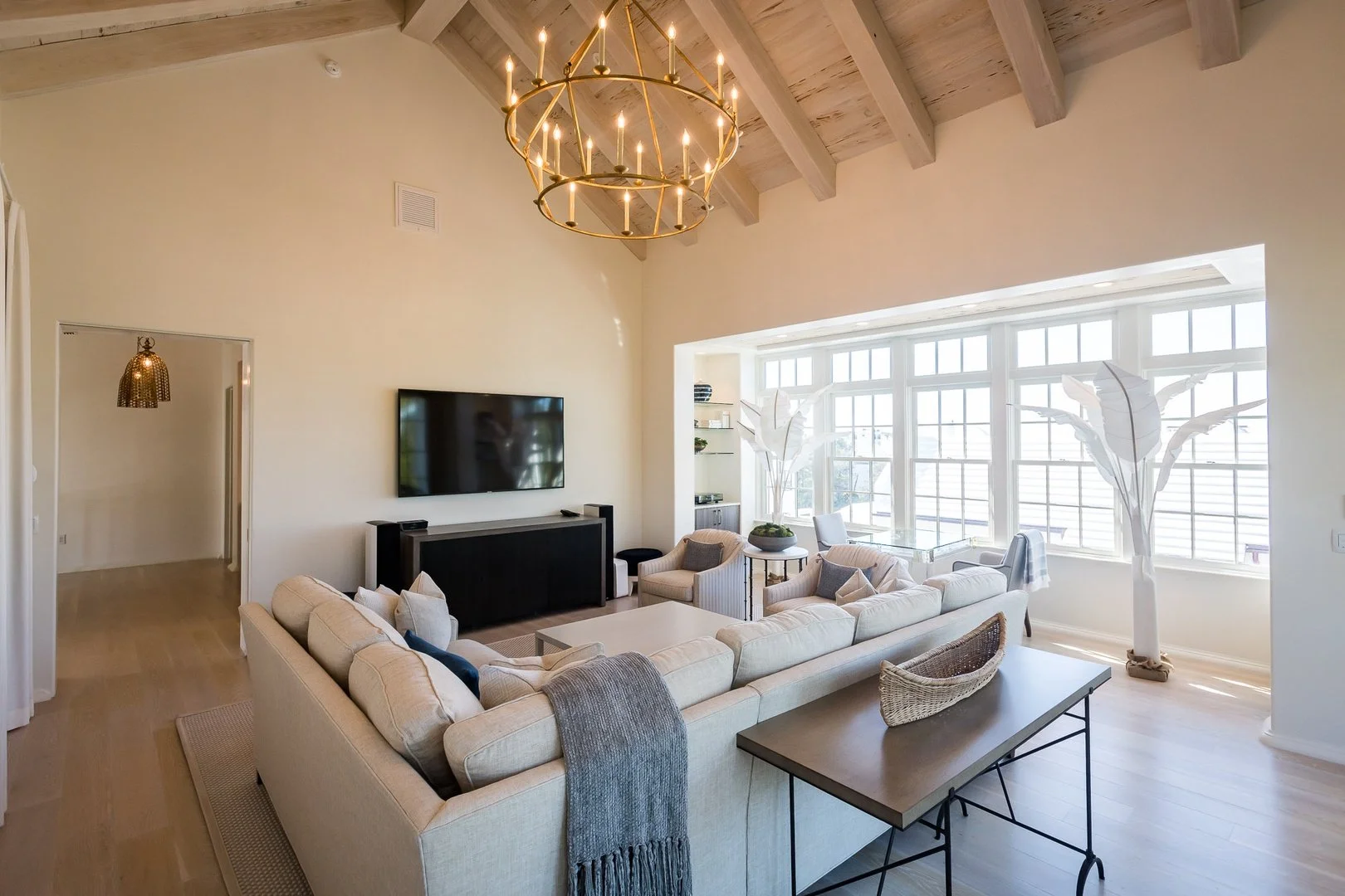 Living room with beige sectional sofa, large TV on wall, chandelier, white interior walls, big windows with blinds, decorative plants, side tables, and a wooden coffee table.