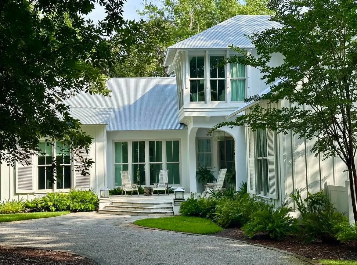 A white two-story house with a blue metal roof, large windows, and a small front porch with steps. The house is surrounded by green trees and bushes, and there are chairs on the porch.
