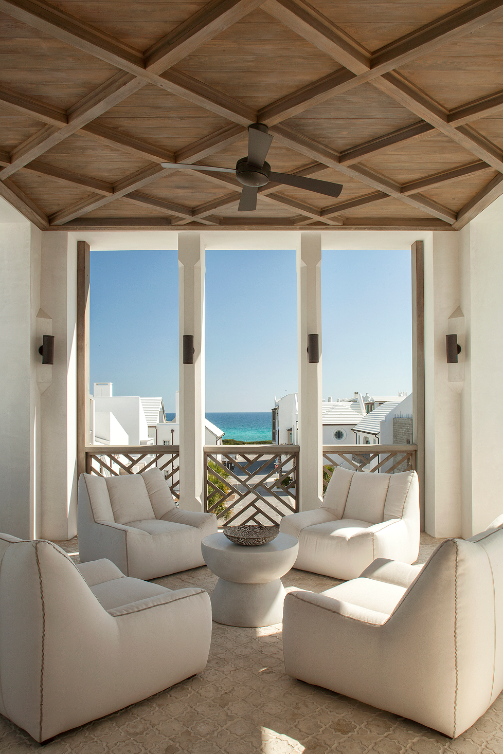 Beachside patio with four white cushioned chairs arranged around a white round table. Open view to white modern houses and the ocean under a clear blue sky, with a wooden ceiling fan overhead.