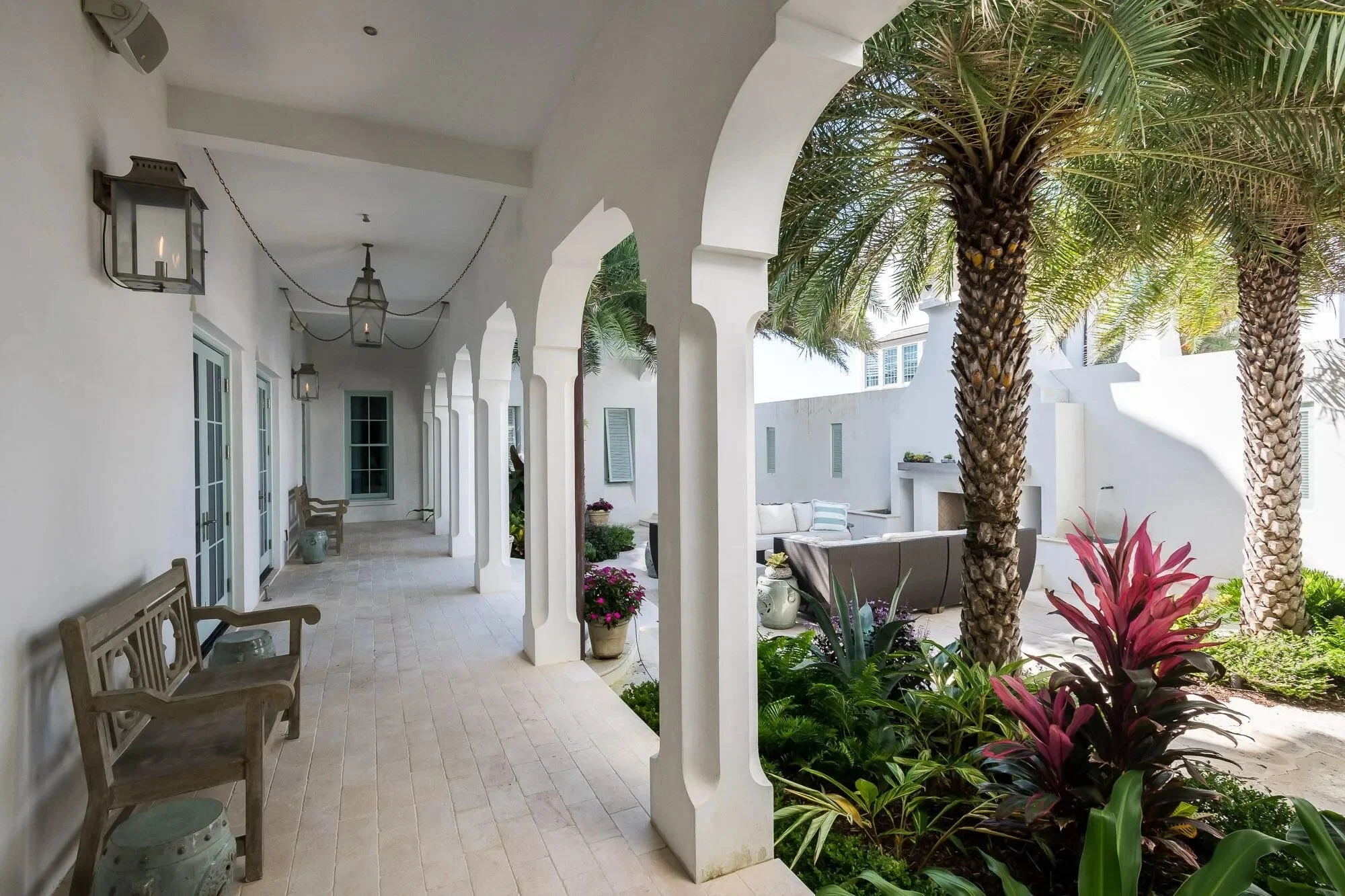 A porch with white walls and arches, wooden benches, and outdoor lighting fixtures; decorated with potted plants and greenery, with palm trees and outdoor seating visible in the background.