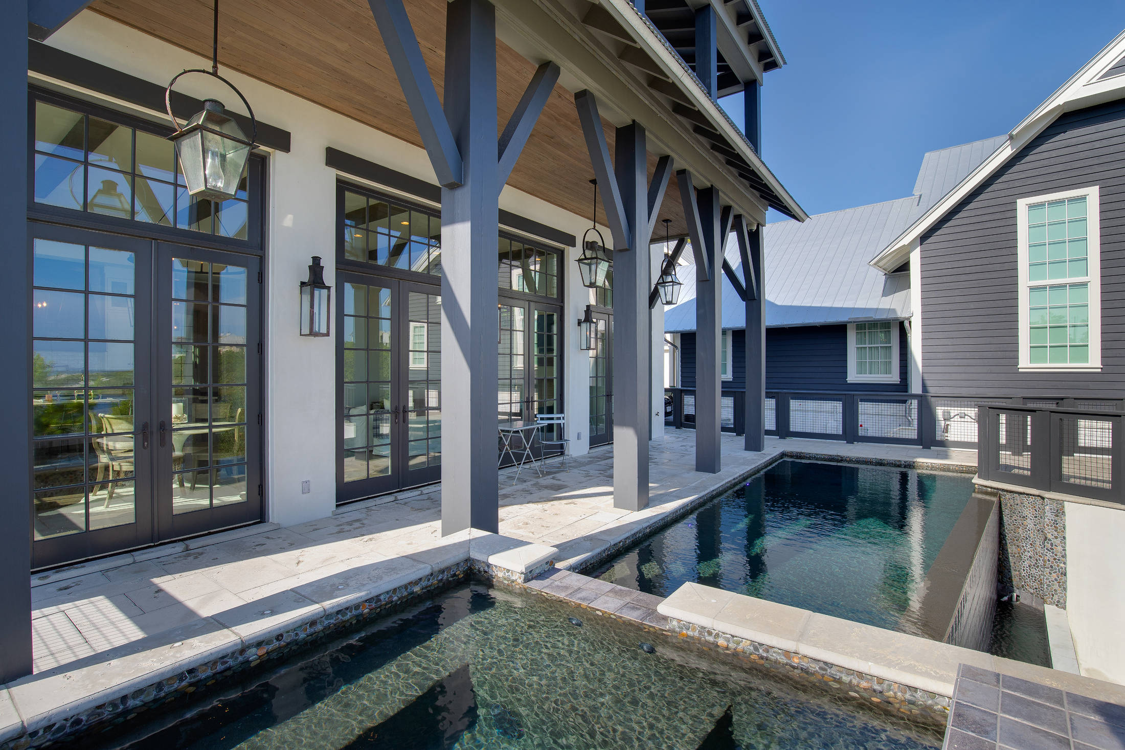 A modern backyard with a small black swimming pool, French doors with grid windows, outdoor pendant lanterns, and a covered patio with wooden beams in the daytime.