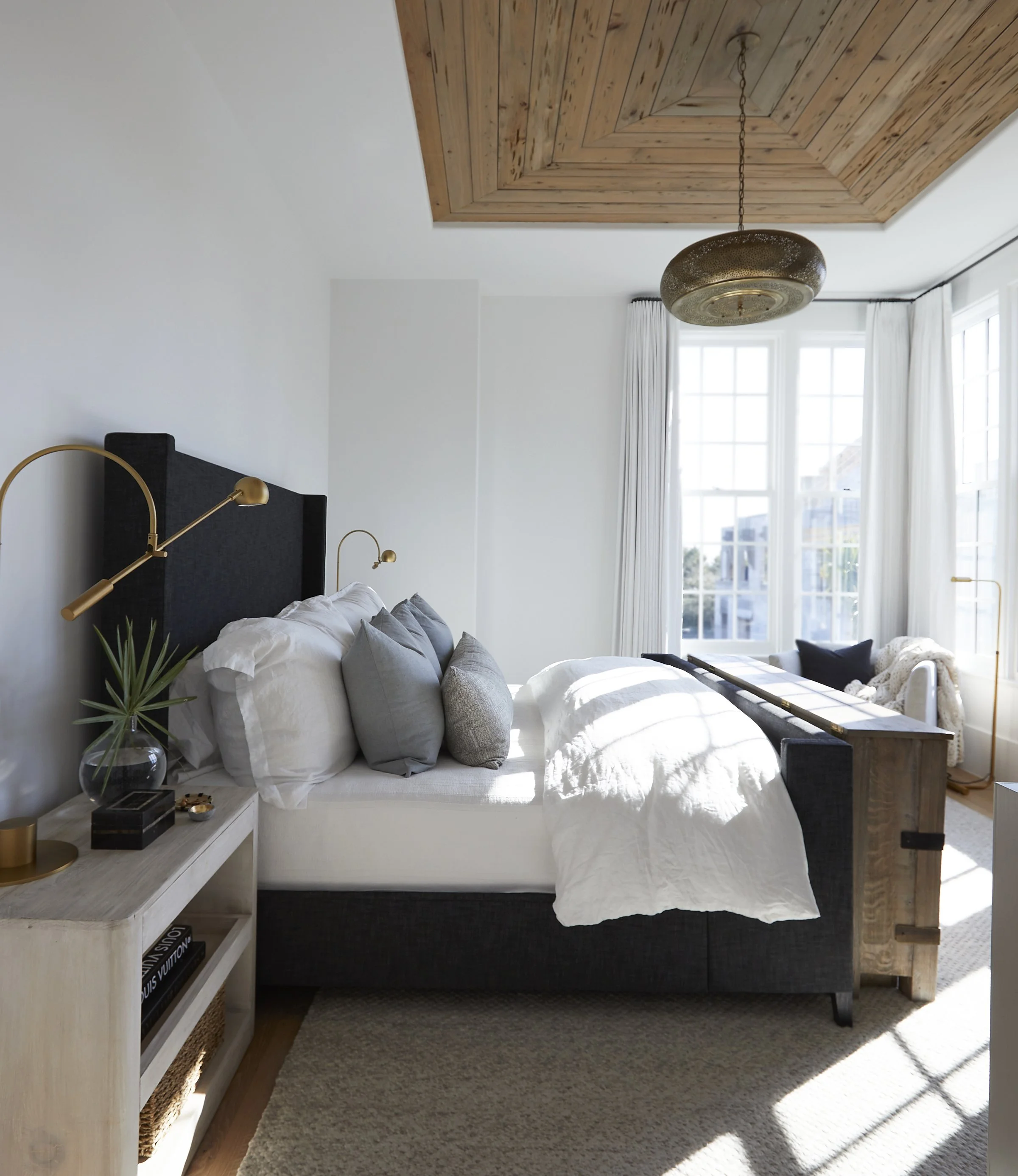 A bright bedroom with a bed, multiple pillows, and a beige blanket. A large window with white curtains allows sunlight to fill the room. There's a white nightstand with decor items and a potted plant on it, and a beige carpet on the floor. The ceilin